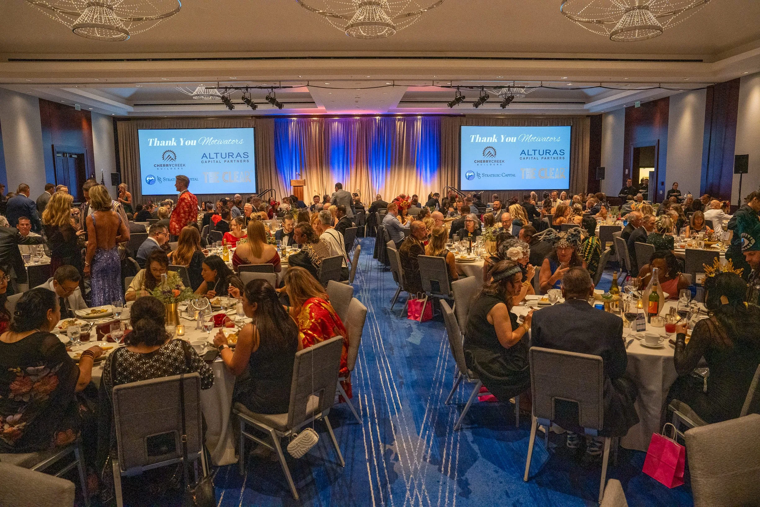 A large banquet hall filled with round tables of people attending a formal event. The stage at the front displays a blue screen with the words 'Thank You Motivators' and logos of various sponsors. The hall is decorated with elegant lighting and drapes.