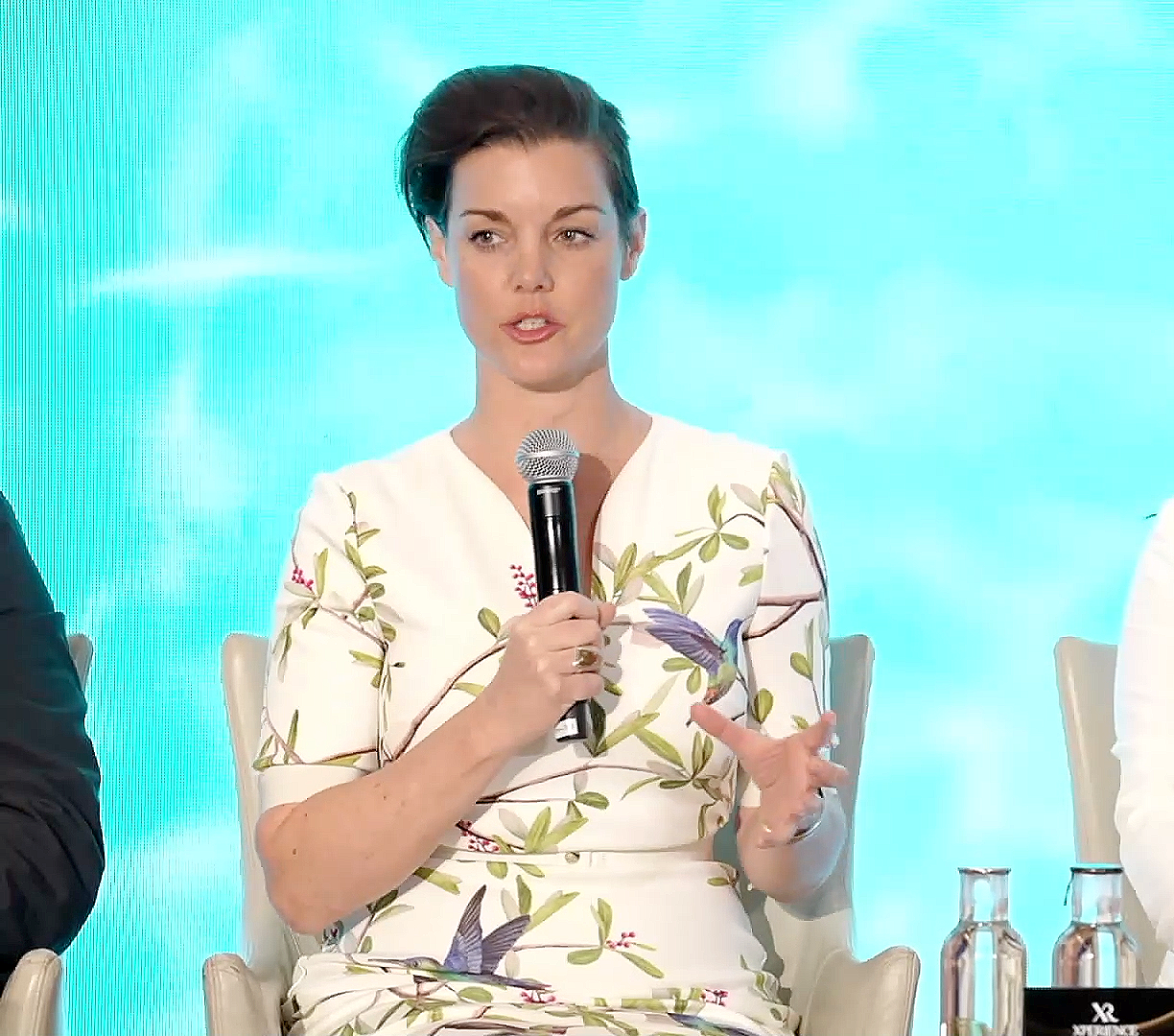 Woman with short dark hair holding a microphone speaking at an event, wearing a white dress with a bird and leaf pattern, sitting on a white chair, with a blue background.