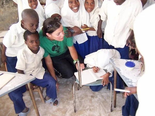 A group of children in school uniforms and a woman in casual clothes gathered around a table in a classroom, with some children seated and others standing, engaging with materials on the table.
