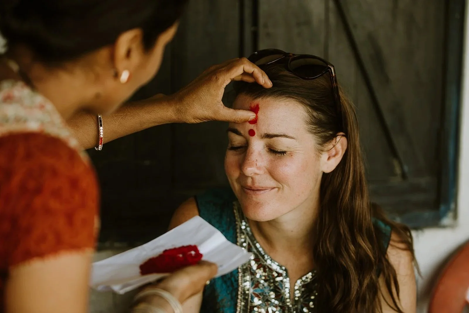A woman with a bindi on her forehead, sitting with eyes closed, as another woman applies red sindoor (vermilion) to her parting hair, during a traditional ceremony.