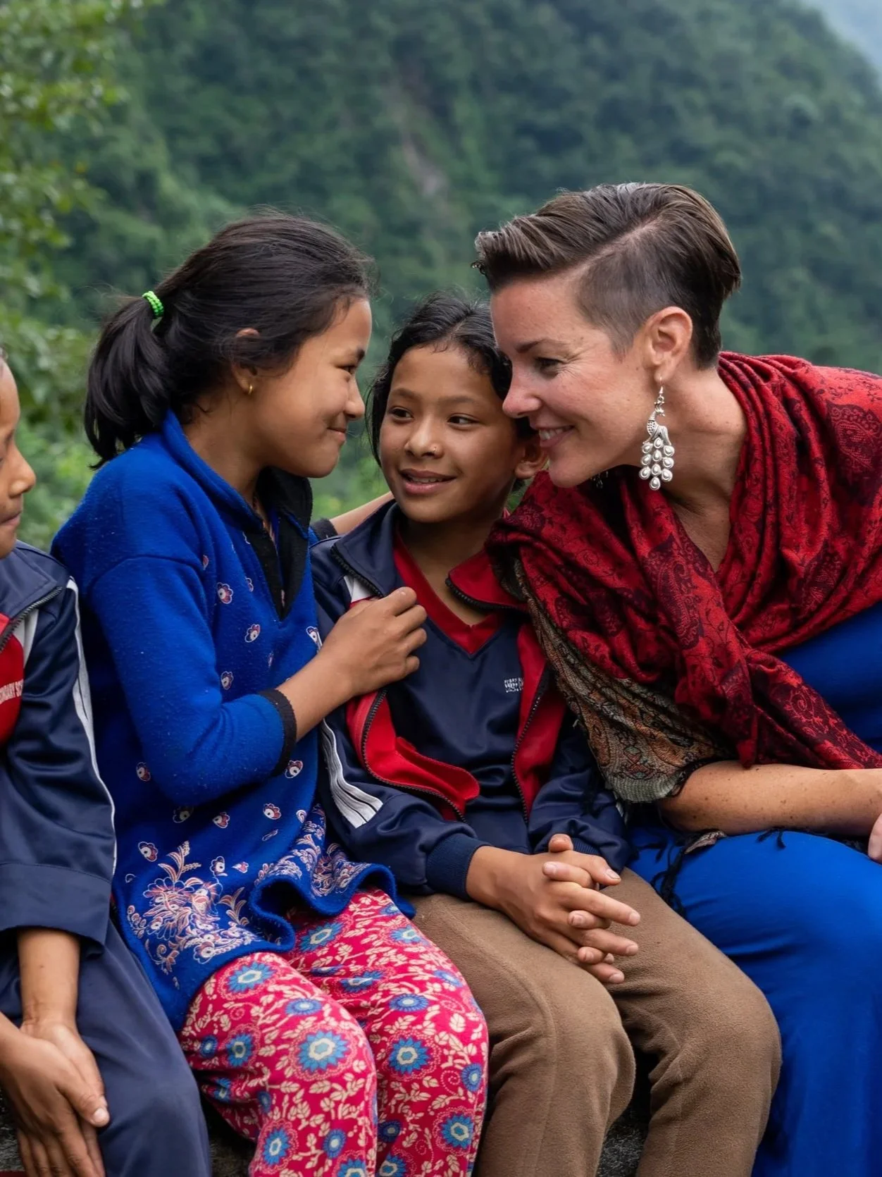 A woman with short hair and earrings smiling and leaning towards two children, a girl and a boy, as they sit outdoors with a green, forested background. The girl is in a blue jacket and the boy in a red and blue jacket, all smiling and engaging closely.