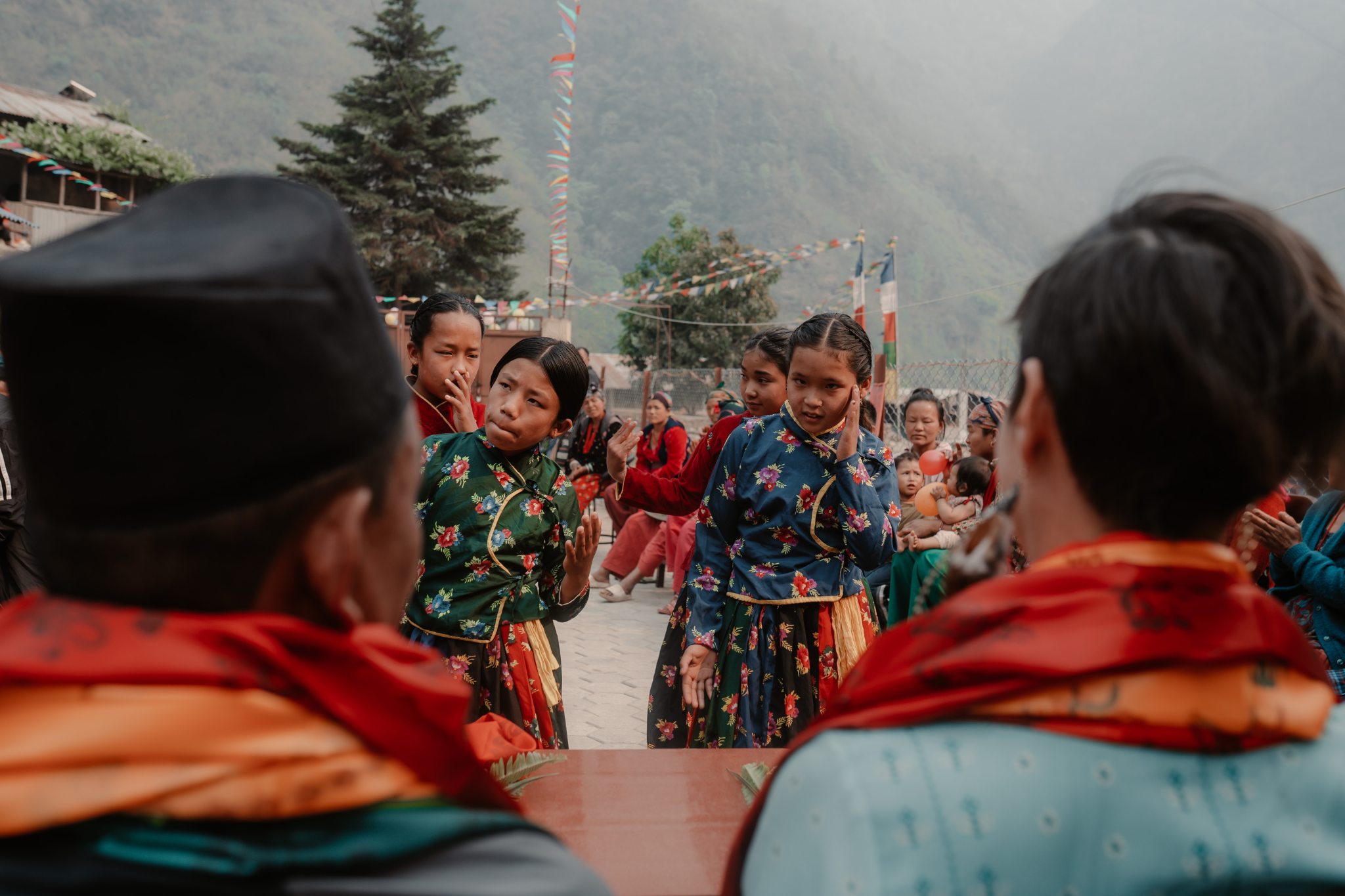 Group of young women in colorful floral traditional dresses dancing in an outdoor setting during a cultural event with onlookers, flags, and mountains in the background.