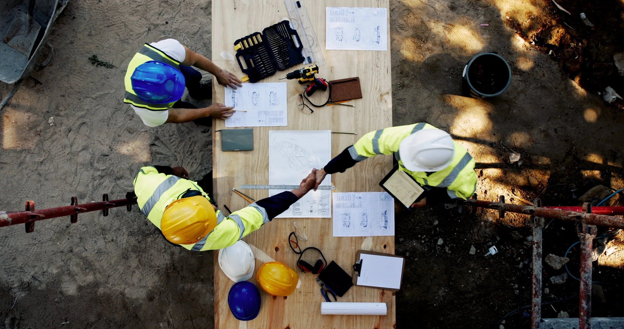 Three construction workers wearing safety helmets and reflective vests shake hands over a wooden table at a construction site. The table has blueprints, tools, and safety equipment. The site has dirt and construction materials around.