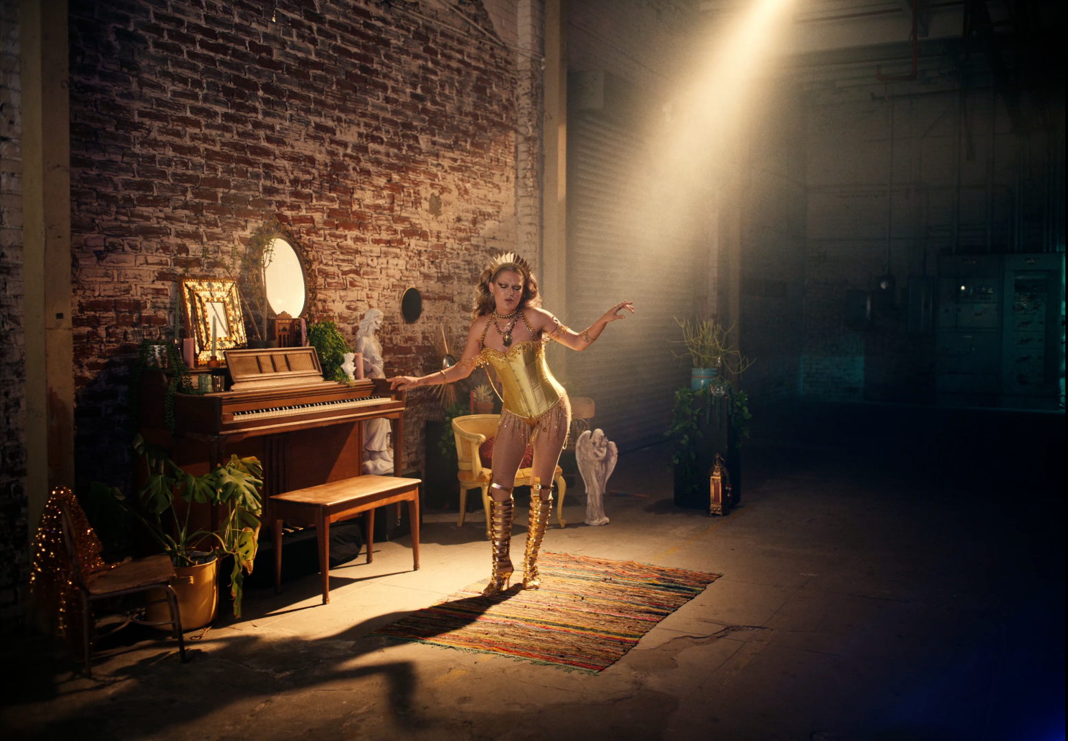 A woman dancing in an industrial-style room with exposed brick walls, lit by a single spotlight. She wears a gold corset, sheer pants, and tall gold boots, with jewelry and a crown. The room contains a vintage piano, mirrors, plants, sculptures, and a colorful rug.