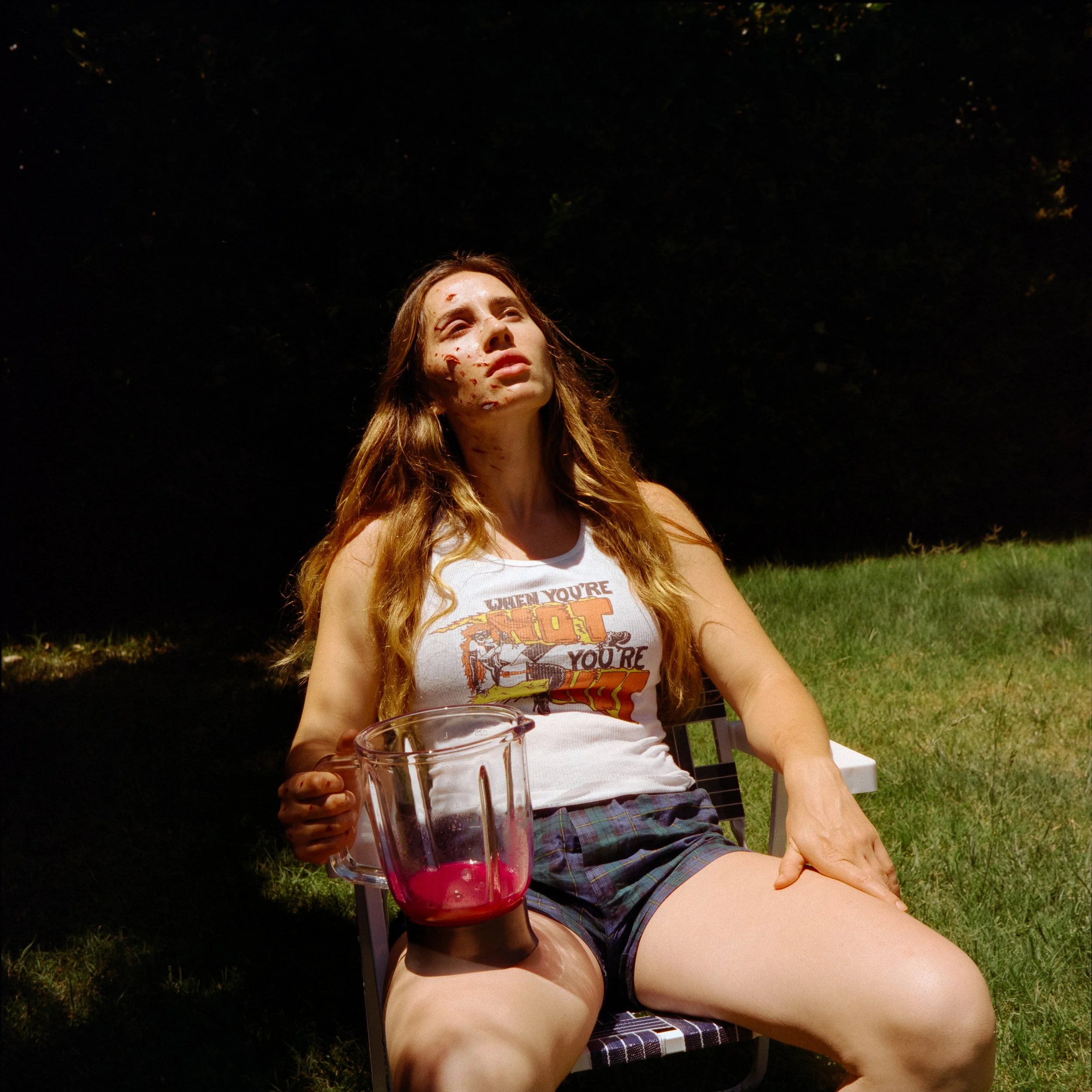 A woman with long, wavy red hair sitting outdoors on a lawn chair, covered in dirt or mud, with a muddy face, wearing a white sleeveless top and plaid shorts, holding a half-full pitcher of pink liquid, looking upwards with a distressed expression.
