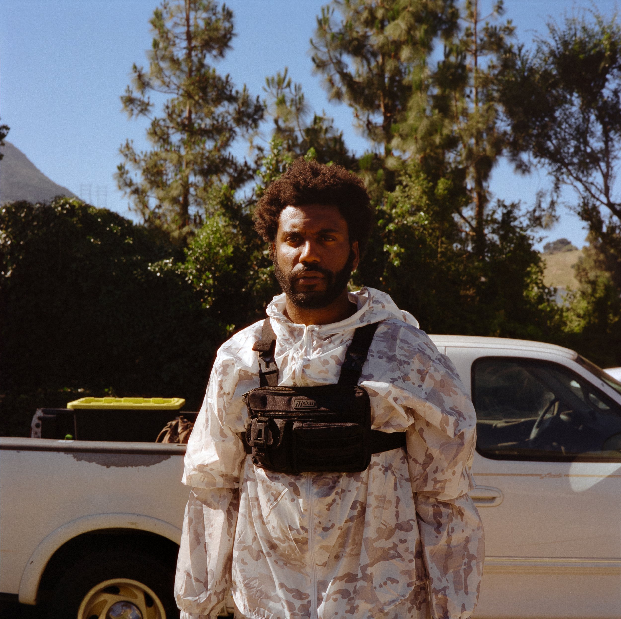 A man with curly hair and a beard wearing a white camouflage jacket and black chest pouch, standing outdoors with trees and a white pickup truck in the background.