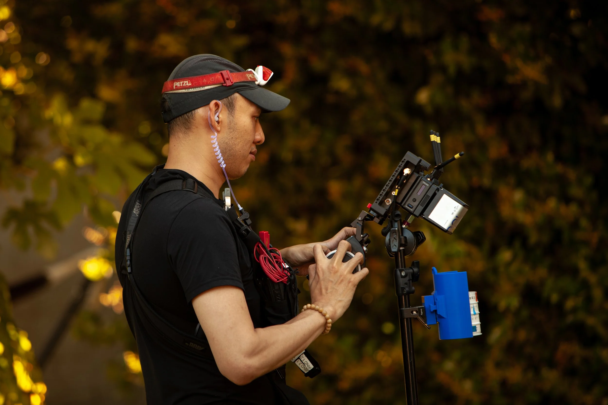 A man wearing a black cap and t-shirt operating a camera mounted on a tripod, with a blue control box attached, outdoors during the evening with fall foliage in the background.