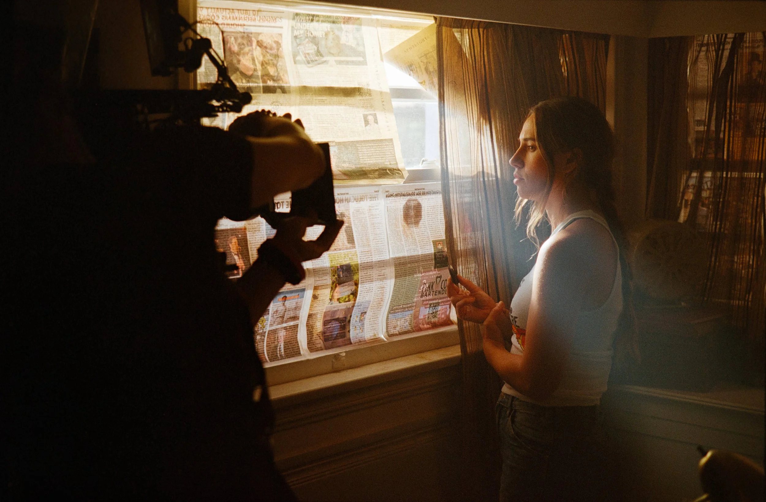 A young woman with shoulder-length hair stands in front of a window covered with newspapers, holding a small object in her hands, as sunlight streams in through the window. A person in the foreground, partially visible, is adjusting a camera to take her photograph.