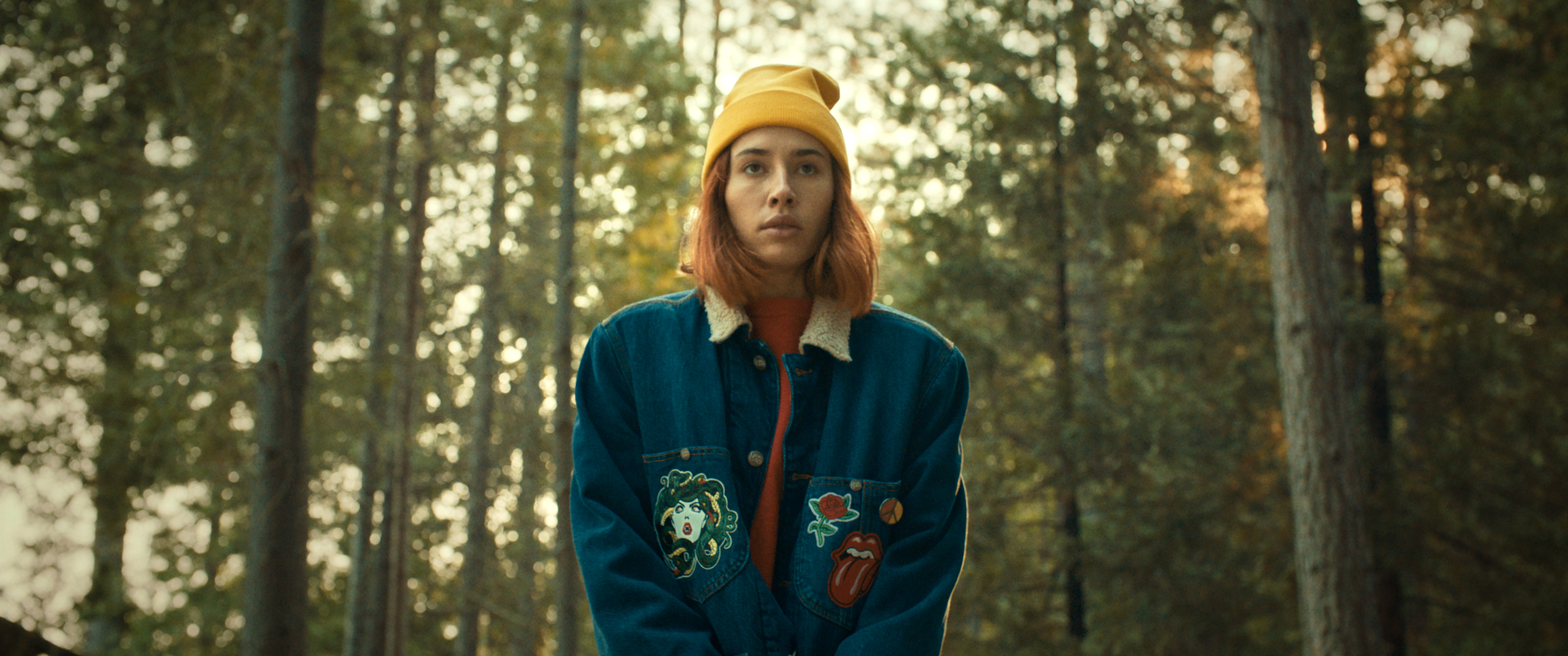 A woman with red hair wearing a yellow beanie and a blue denim jacket with patches, standing in a forest during sunset.