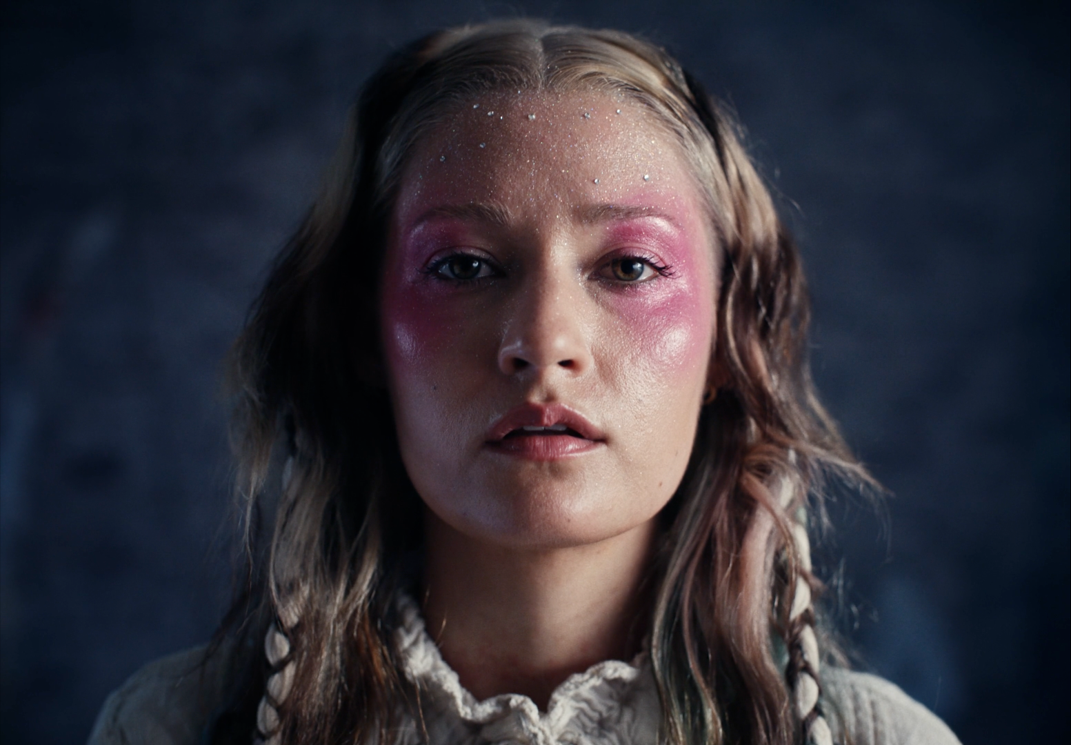 A close-up of a young woman with wet, wavy hair and pink makeup on her face, looking directly at the camera with an intense expression.