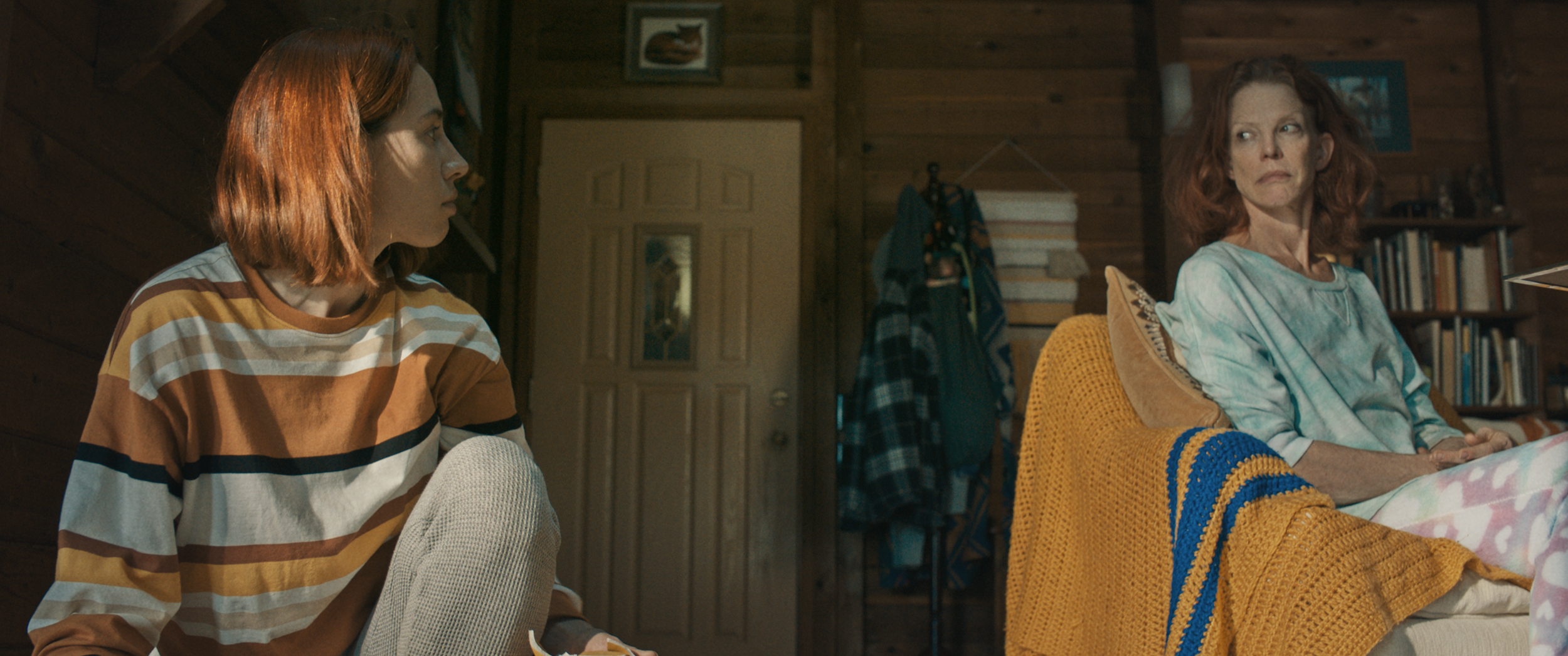 A young woman with red hair wearing a striped brown, white, black, and yellow shirt sits on the floor looking to the side. An older woman with red hair wearing a light tie-dye shirt sits on a couch with patterned blankets, looking sternly to the side. The room has wooden walls, bookshelves, and a framed picture of a fox on the wall.