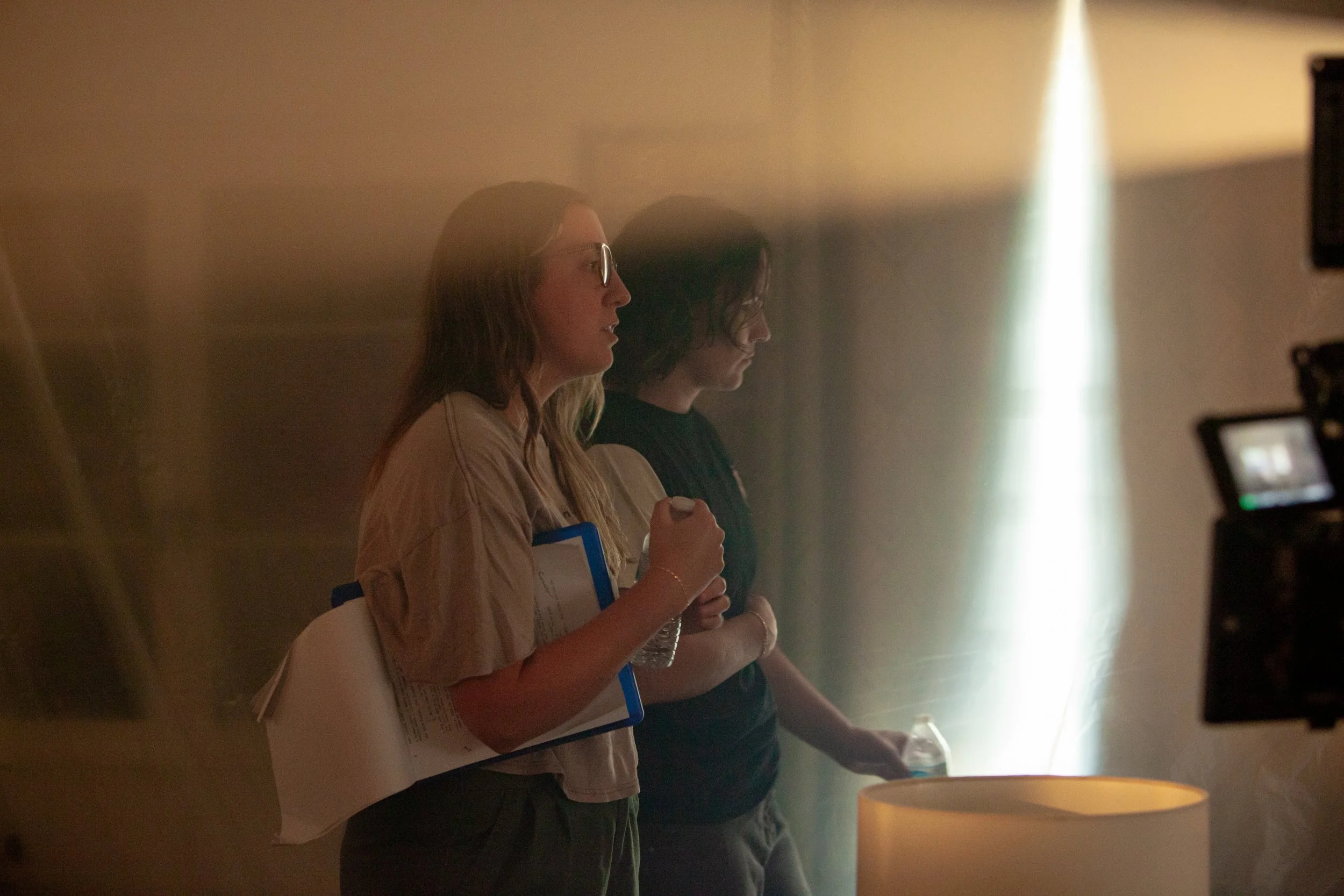 Two young women standing in a softly-lit room, facing to the right, with one holding a water bottle and clipboard, and the other holding a plastic cup, near a table with a lamp.