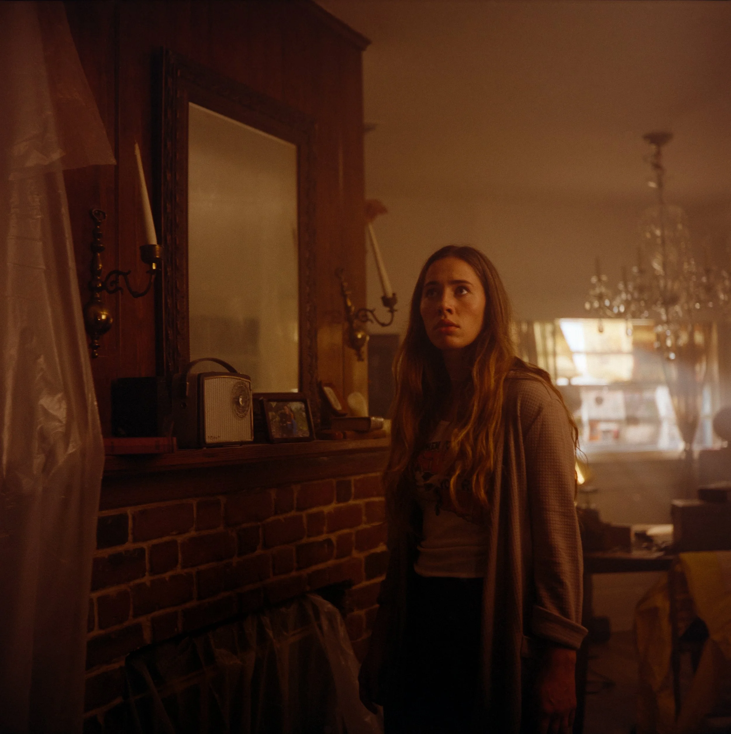 A young woman with long, wavy red hair looks to the side in a dimly lit room with warm, orange lighting. The room has a brick fireplace mantle with picture frames, a vintage radio, and a mirror, along with a chandelier hanging from the ceiling.