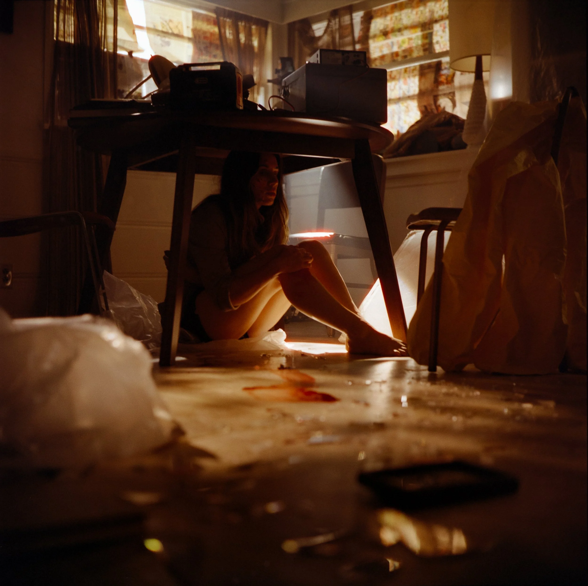 A woman sitting on the floor under a table in a dimly lit room with sunlight coming through a window, surrounded by scattered trash, bottles, and plastic bags.