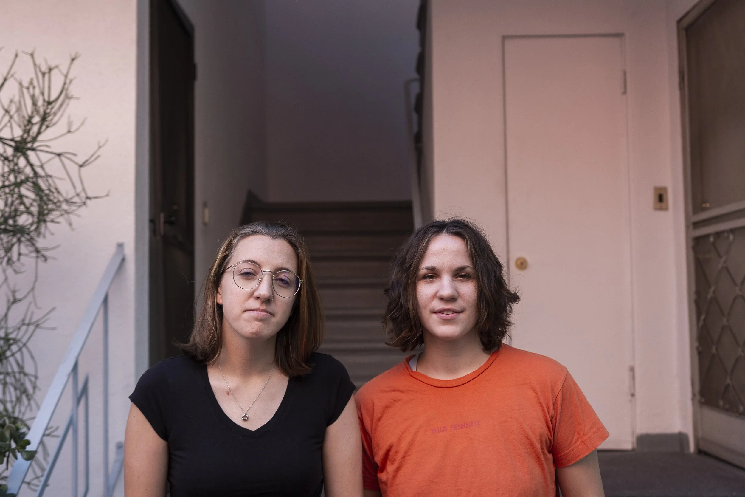 Two young women standing close together outside a building, with a staircase in the background.