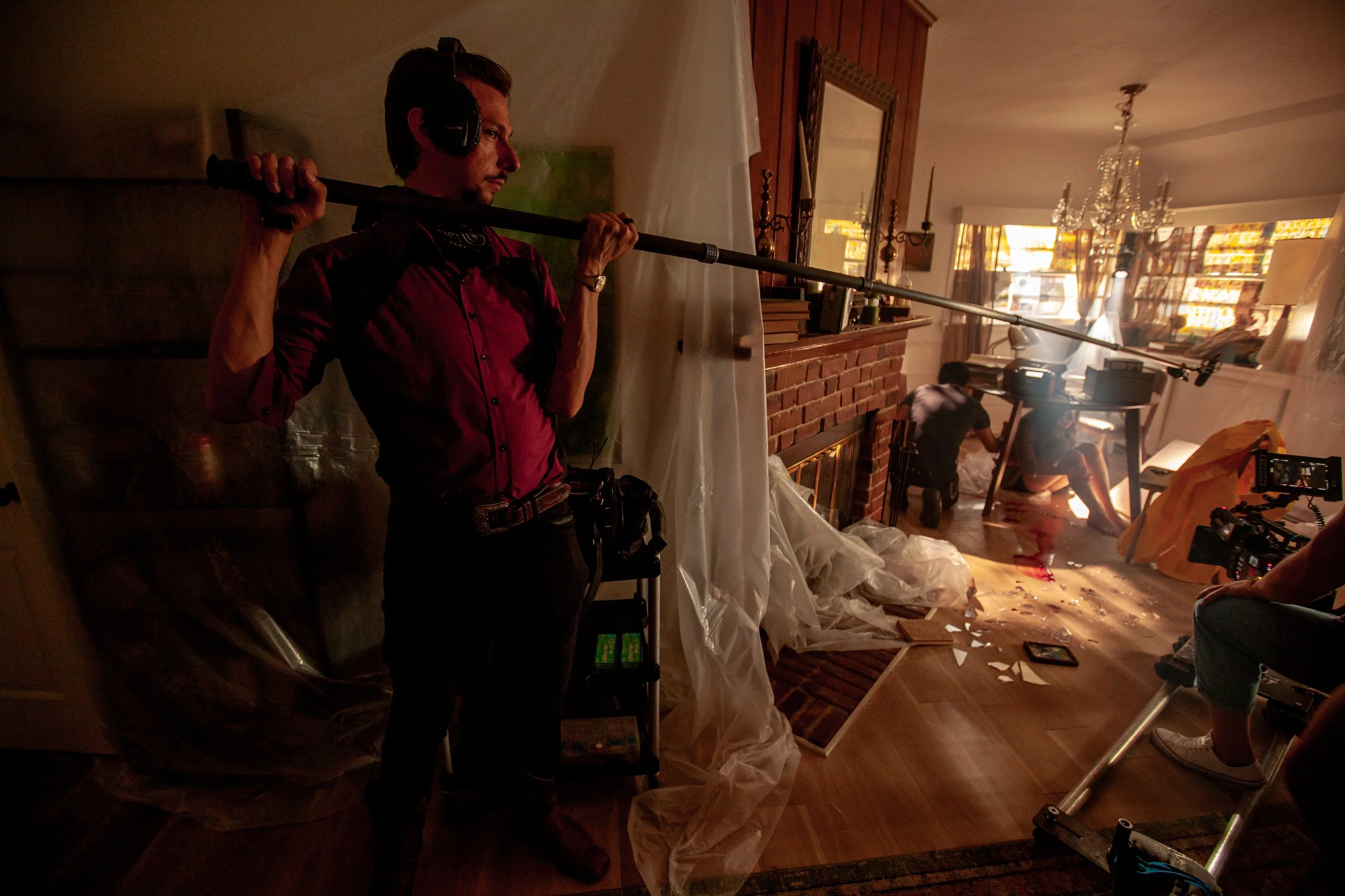 On set, a man is holding a boom microphone over a chaotic scene in a living room, which appears to be a film or TV production with crew members working amidst debris.