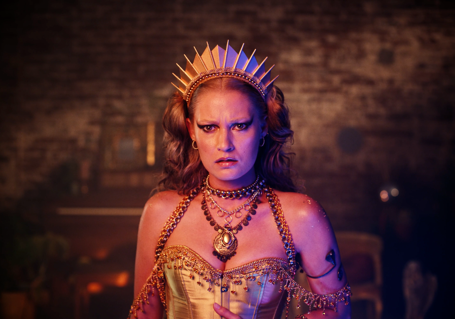A woman with wavy hair wearing a gold crown, multiple layers of jewelry, and a gold corset, standing against a dark, textured background with serious, intense expression.