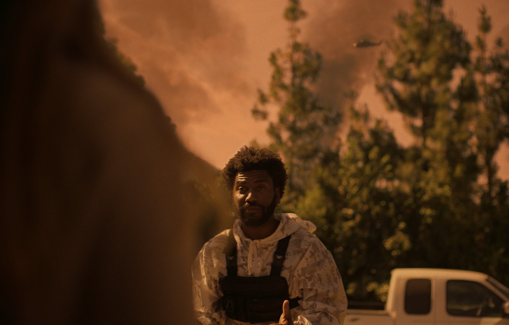 A man with curly hair and a beard wearing a camouflage jacket and a black vest, standing outdoors at night with trees and a vehicle behind him.