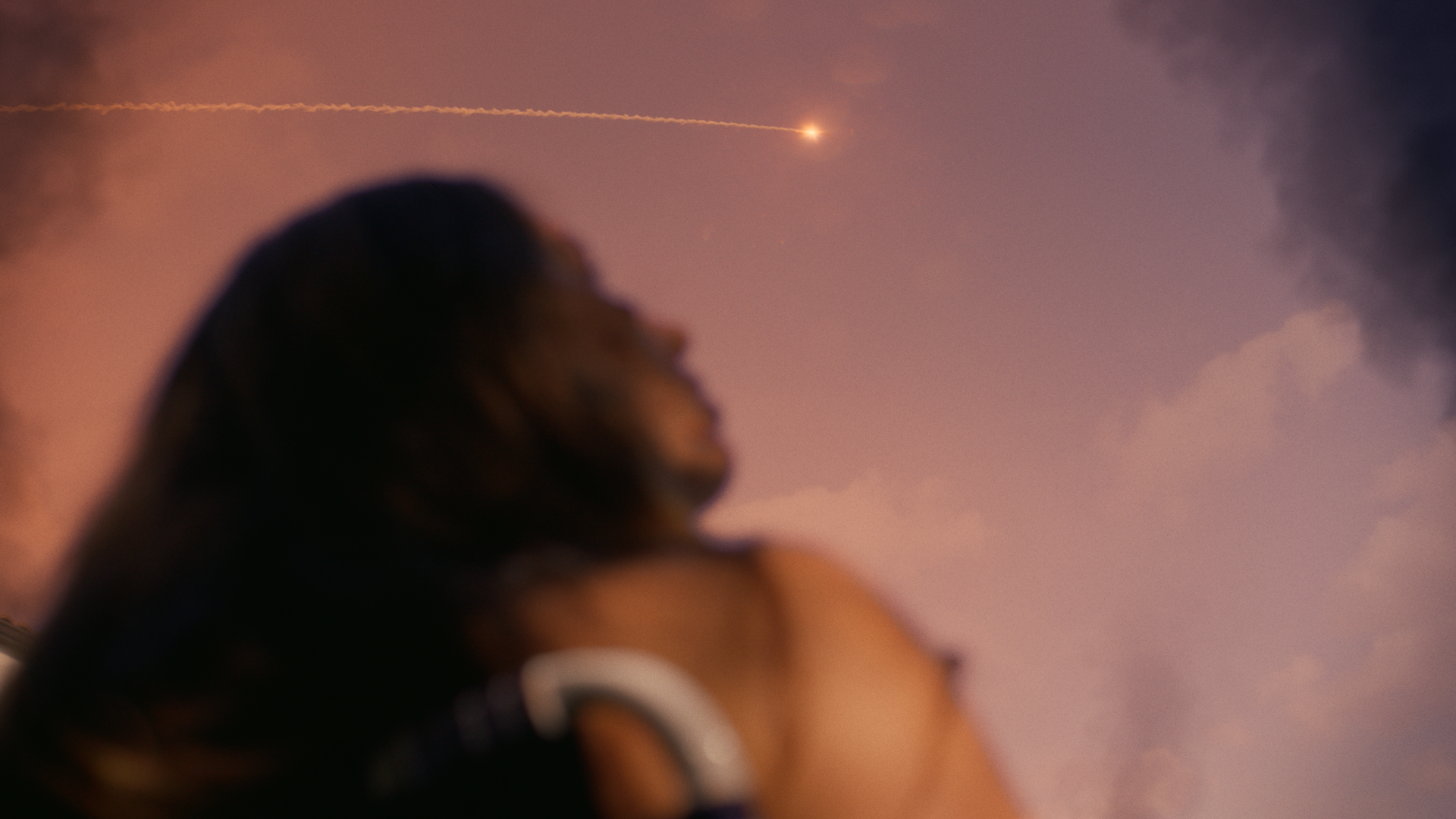 A blurred portrait of a woman looking up at the sky at night, with a bright streak of light or fireball in the sky.