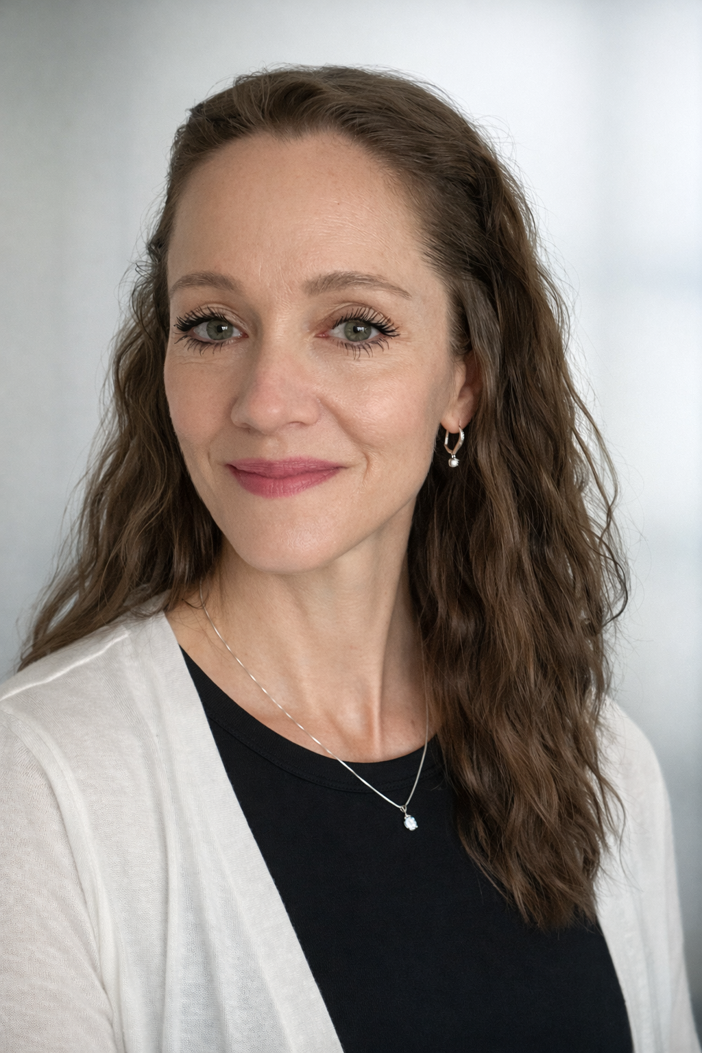 A woman with light skin and curly brown hair smiling slightly, wearing a black top, white cardigan, and jewelry including earrings and a necklace, against a neutral background.
