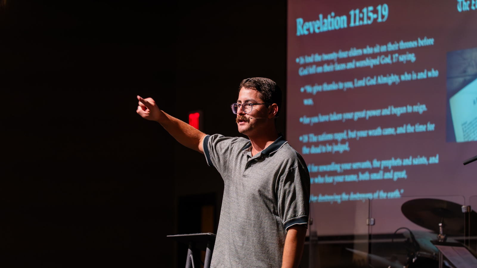 A man wearing glasses and a gray polo shirt is pointing at a projection screen during a presentation, with Bible verses from Revelation 11:15-19 displayed on the screen.
