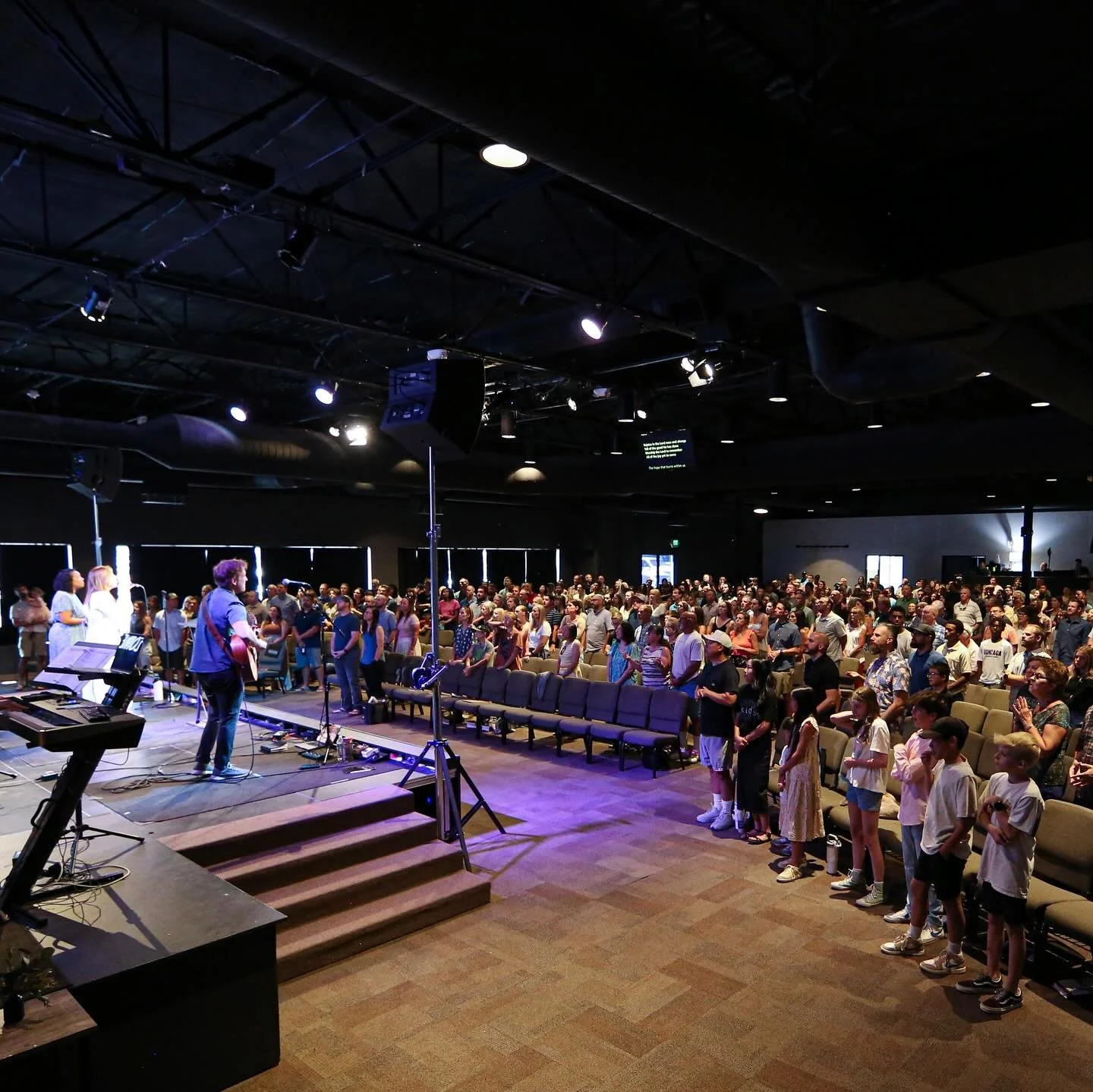 A large indoor gathering with a stage where musicians are performing, and a congregation standing and singing, in a church or concert hall setting.