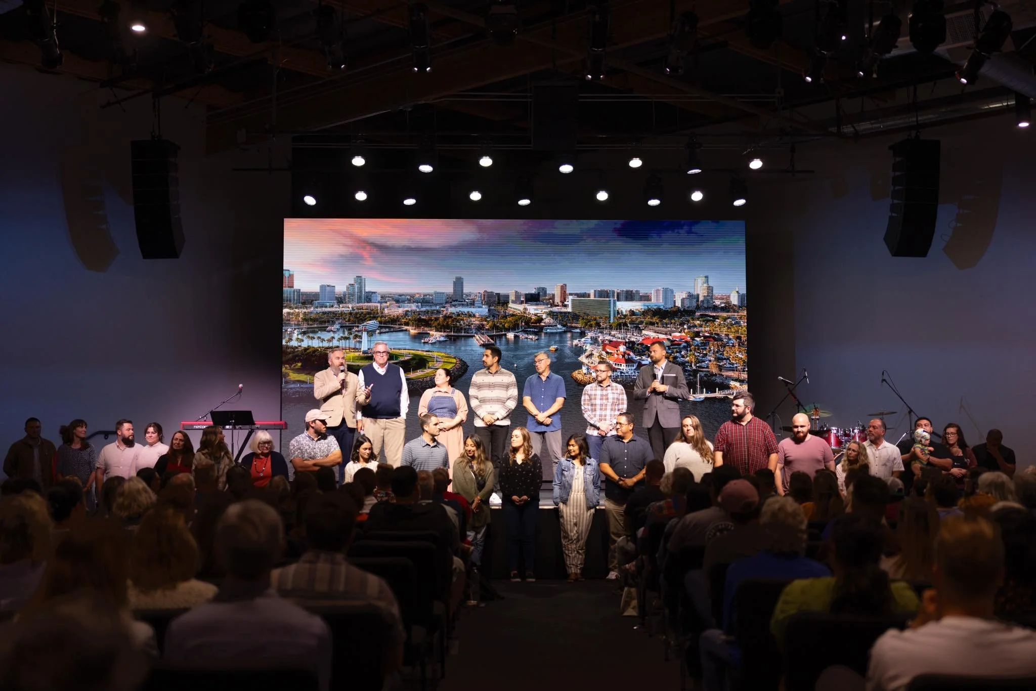 Group of people on stage during a presentation, with a large screen showing a city skyline in the background and an audience watching.