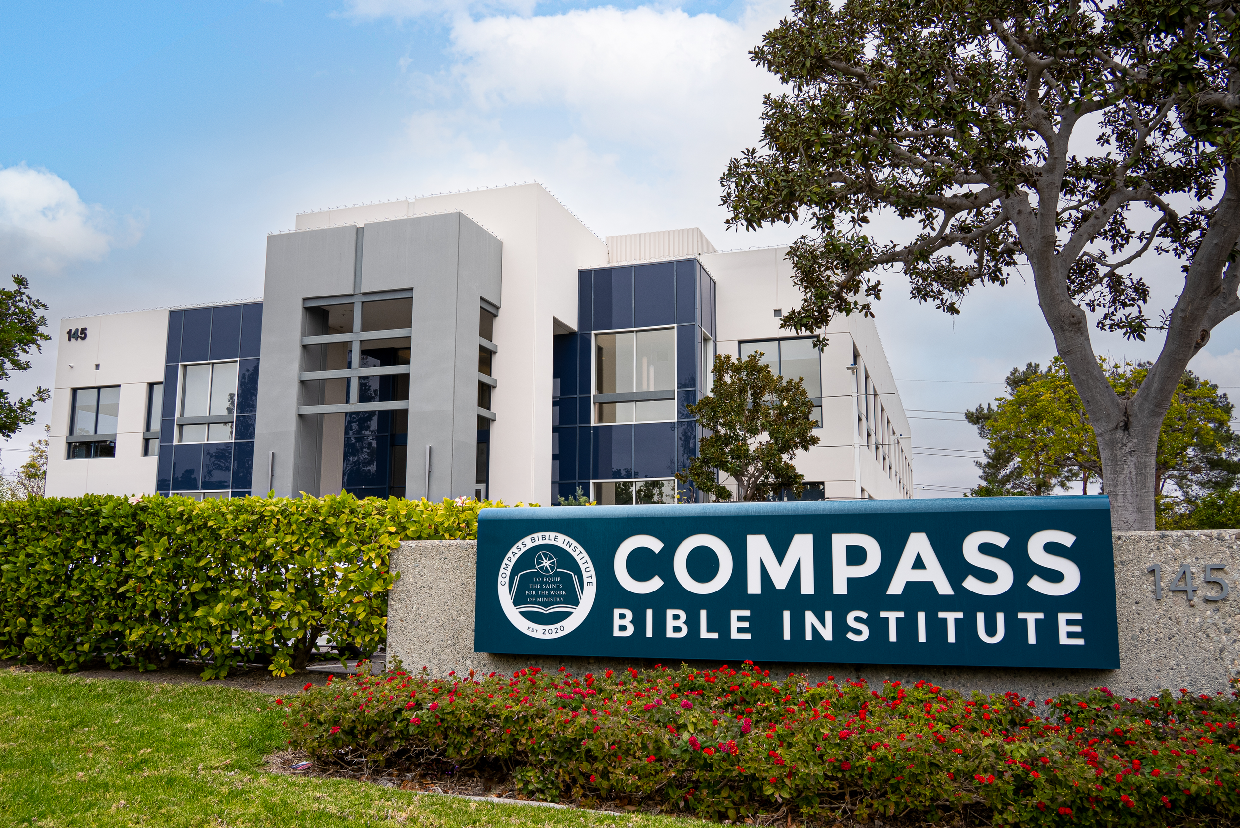 Modern building with a sign for Compass Bible Institute and surrounding landscaping including bushes and trees.