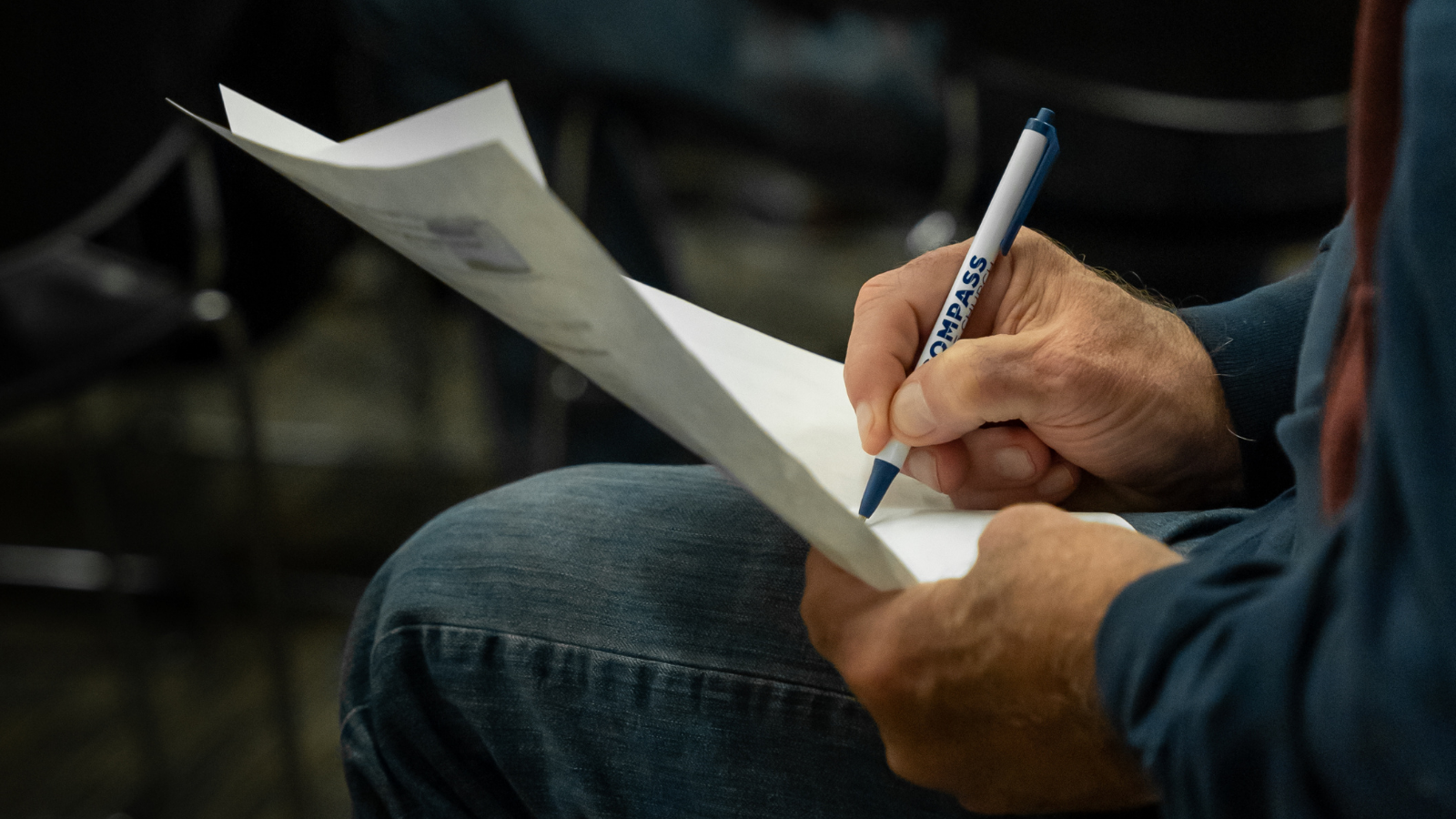 Person writing on a piece of paper with a blue and white pen, seated in a dark room with chairs.