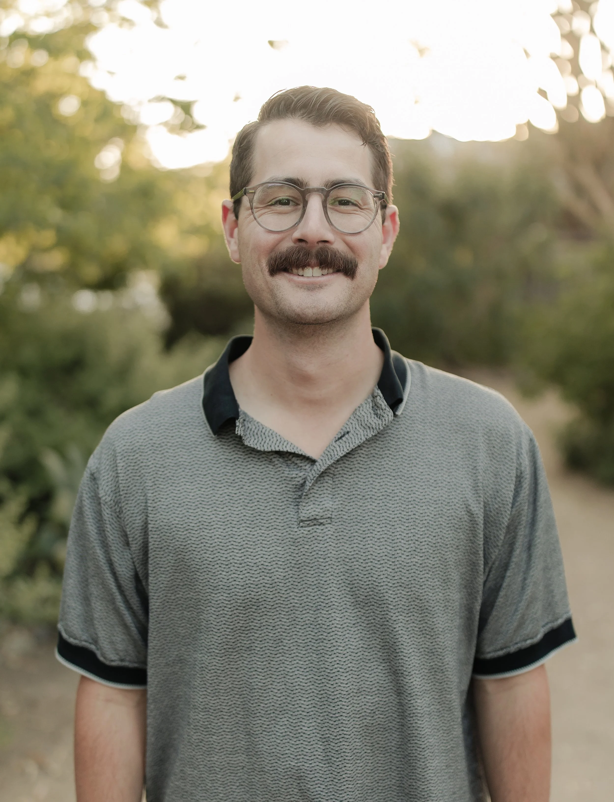 Close-up of a smiling man with glasses and a mustache, outdoors in a park with trees in the background.