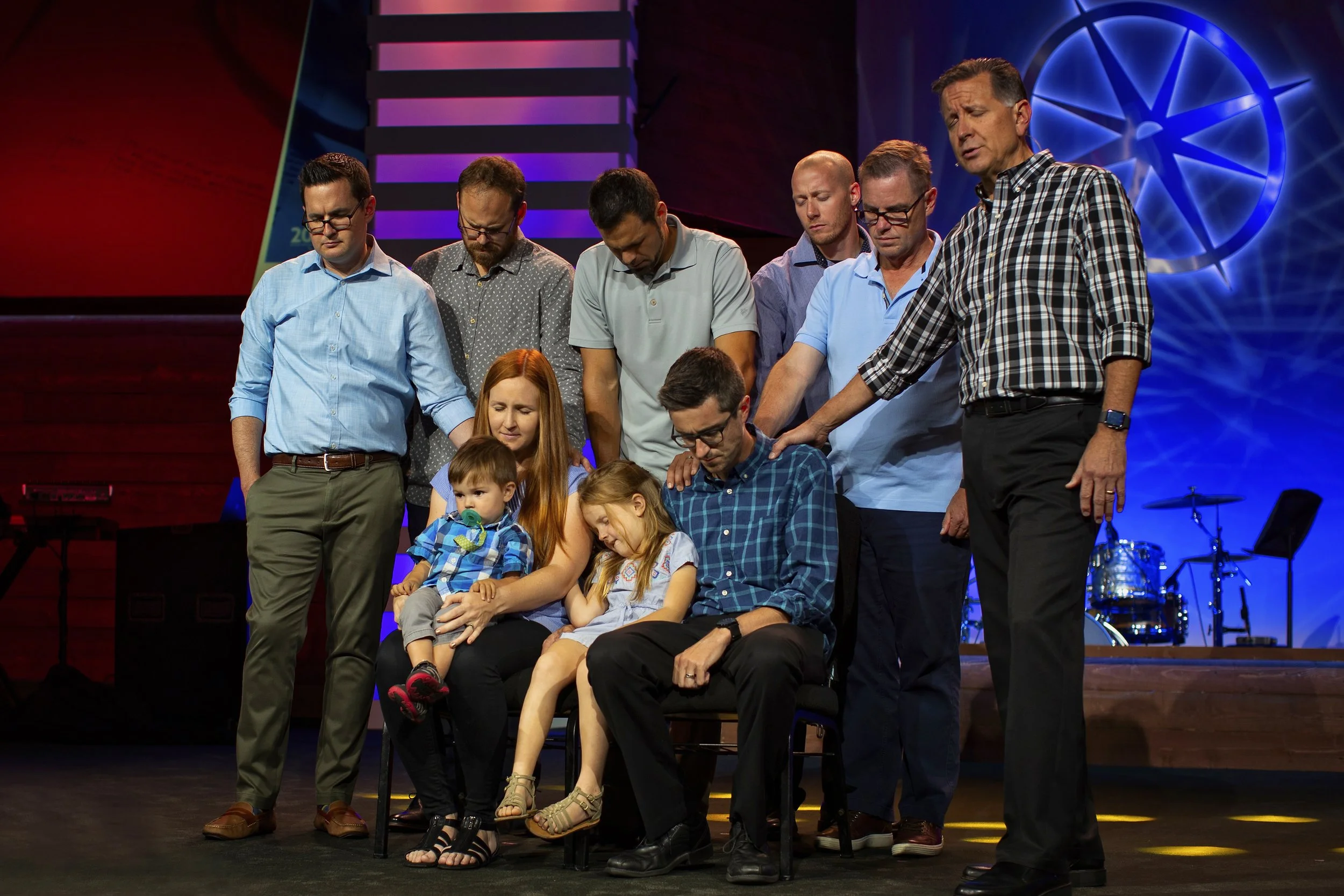 A group of people, including children, participating in a prayer circle on stage with hands on each other's shoulders or heads.