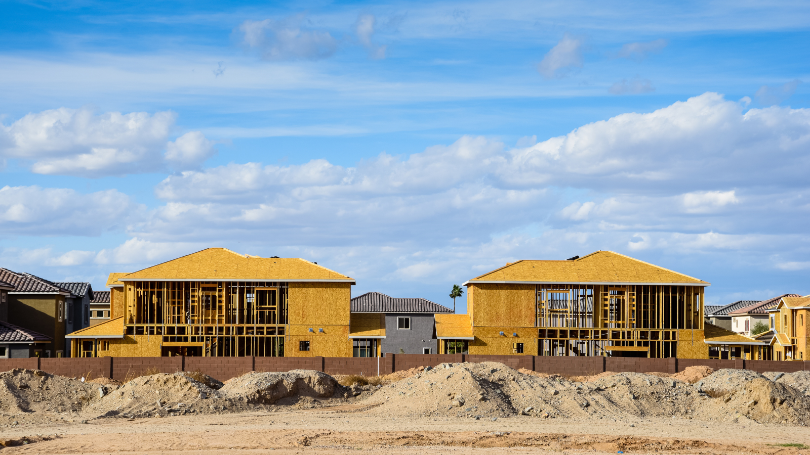 Residential construction site with two houses in framed wood stages, surrounded by other finished houses and a dirt foreground, under a blue sky with clouds.