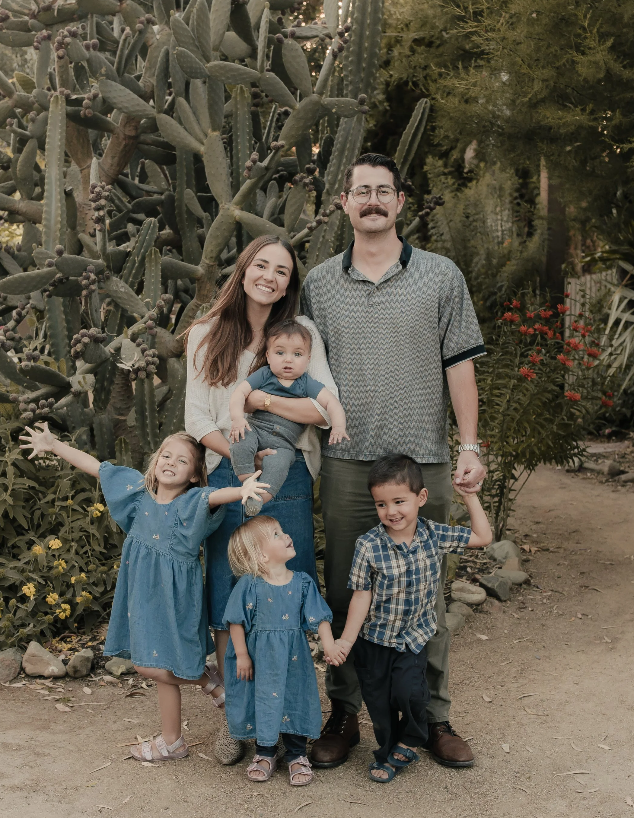A family of four with two young children posing outdoors in front of a large cactus and some green trees. The woman is holding a young child, and they are all smiling or looks happy. The children are wearing blue dresses, and the man and woman are dressed casually.