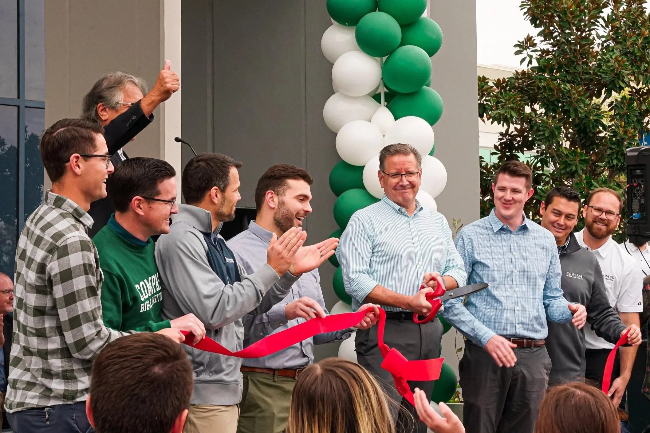 A man in a light blue, striped shirt is holding large red scissors, about to cut a red ribbon at a ribbon-cutting ceremony. Several men stand beside him, clapping and smiling. There is a decorative balloon arch made of green and white balloons behind them, and an audience is visible in the foreground.