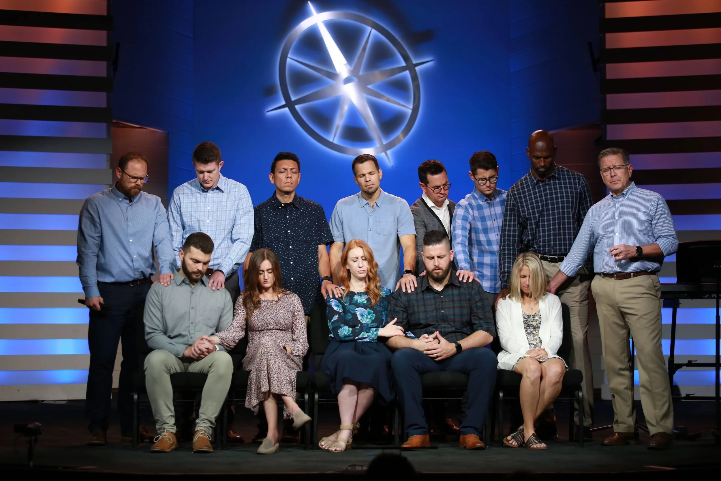 Group of people in a church or conference setting, some kneeling and some standing, with hands placed on shoulders or heads of others, in front of a blue background with illuminated geometric symbol.