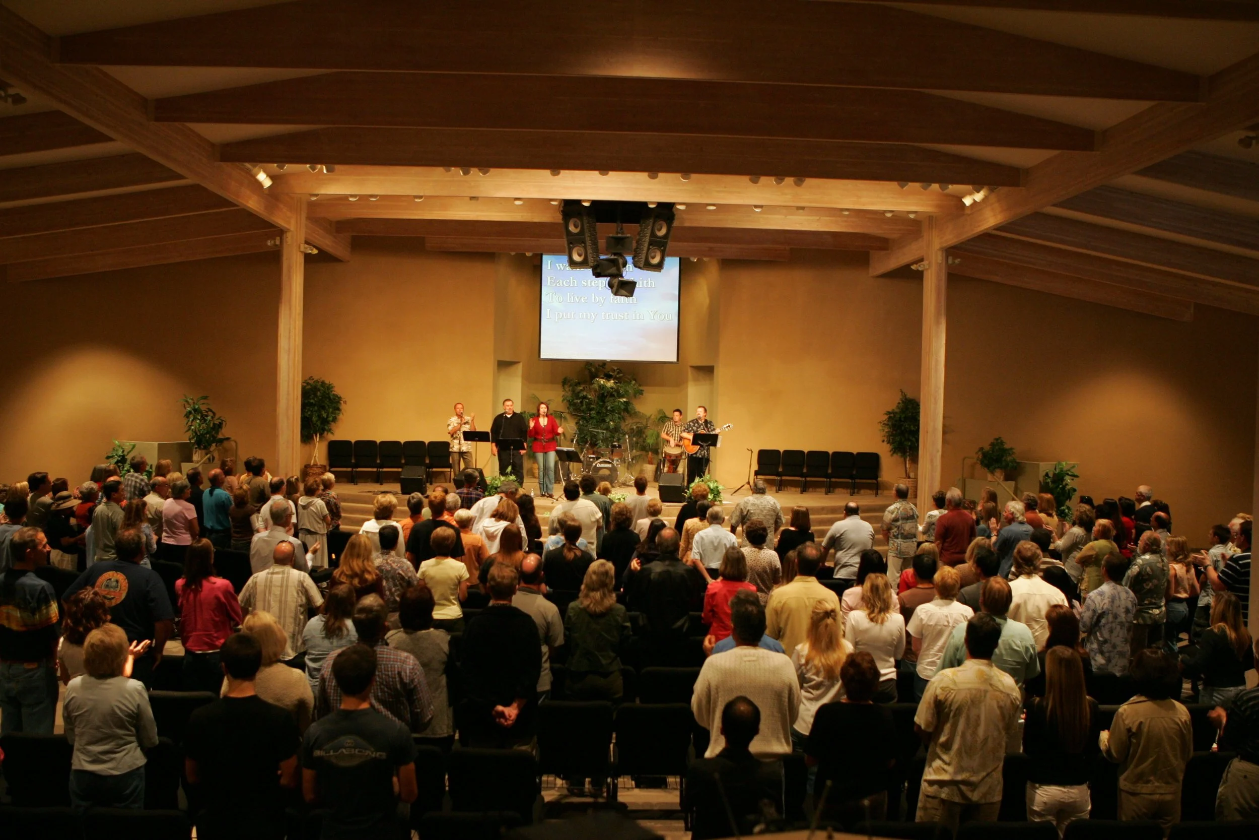 People gathered in a church, participating in a worship service with a band on stage, singing with lyrics displayed on a large screen.