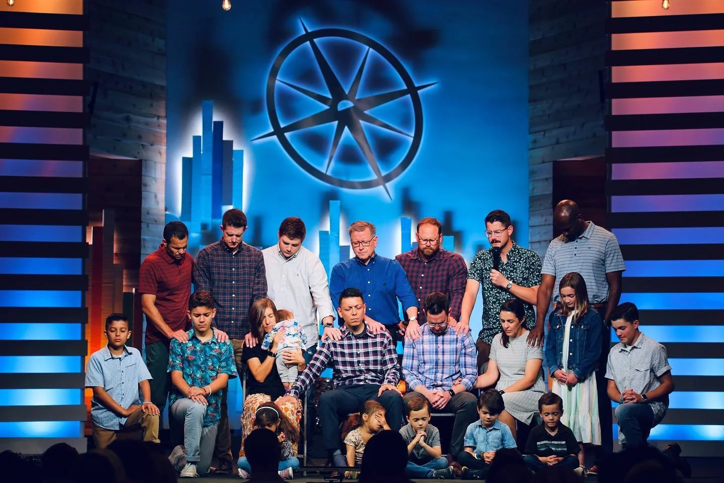 Group of people praying on stage in front of a blue background with a large circle and compass symbol.