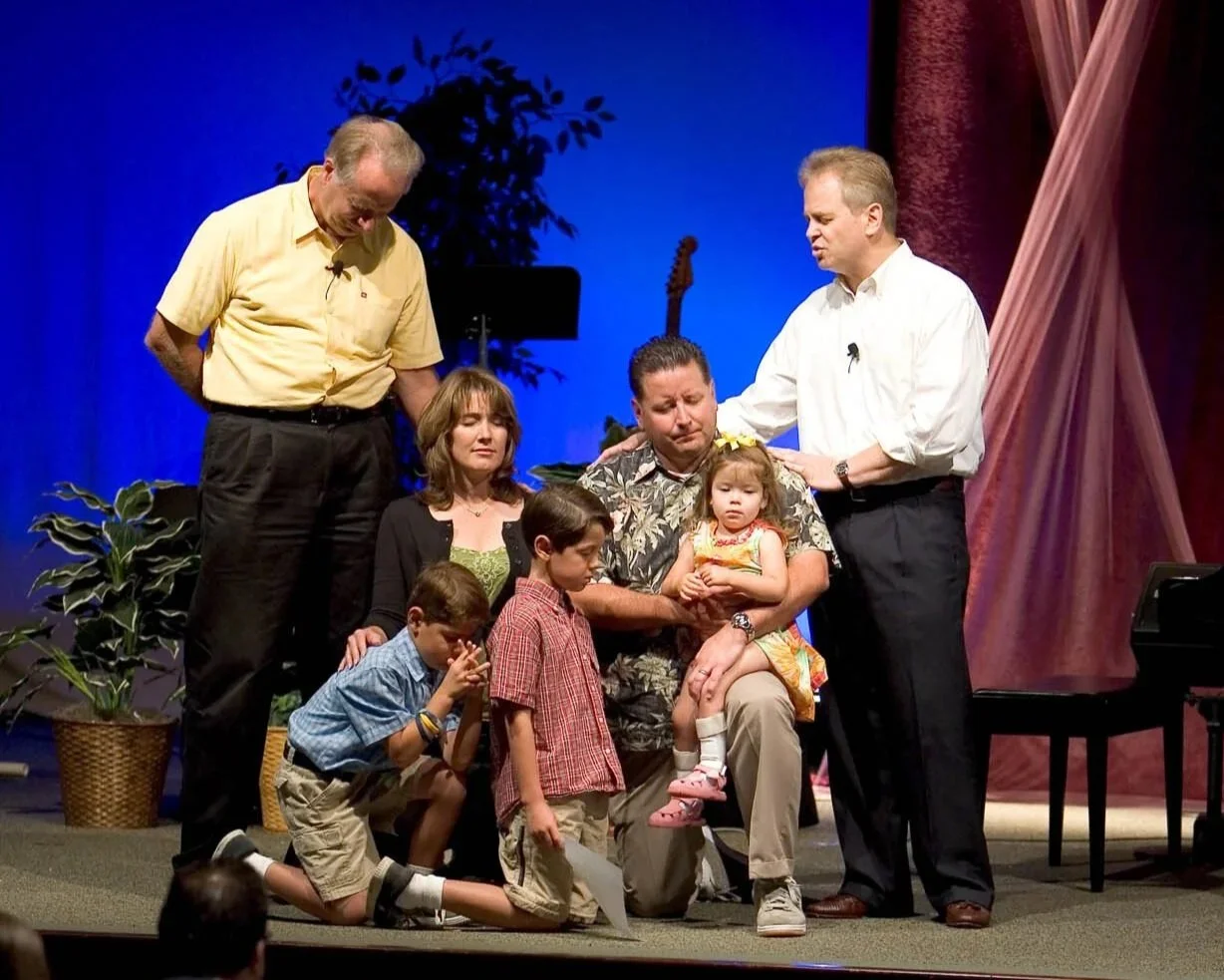 A group of seven people, including children, praying or laying hands on a man who appears distressed, on a stage with plants and a guitar in the background.