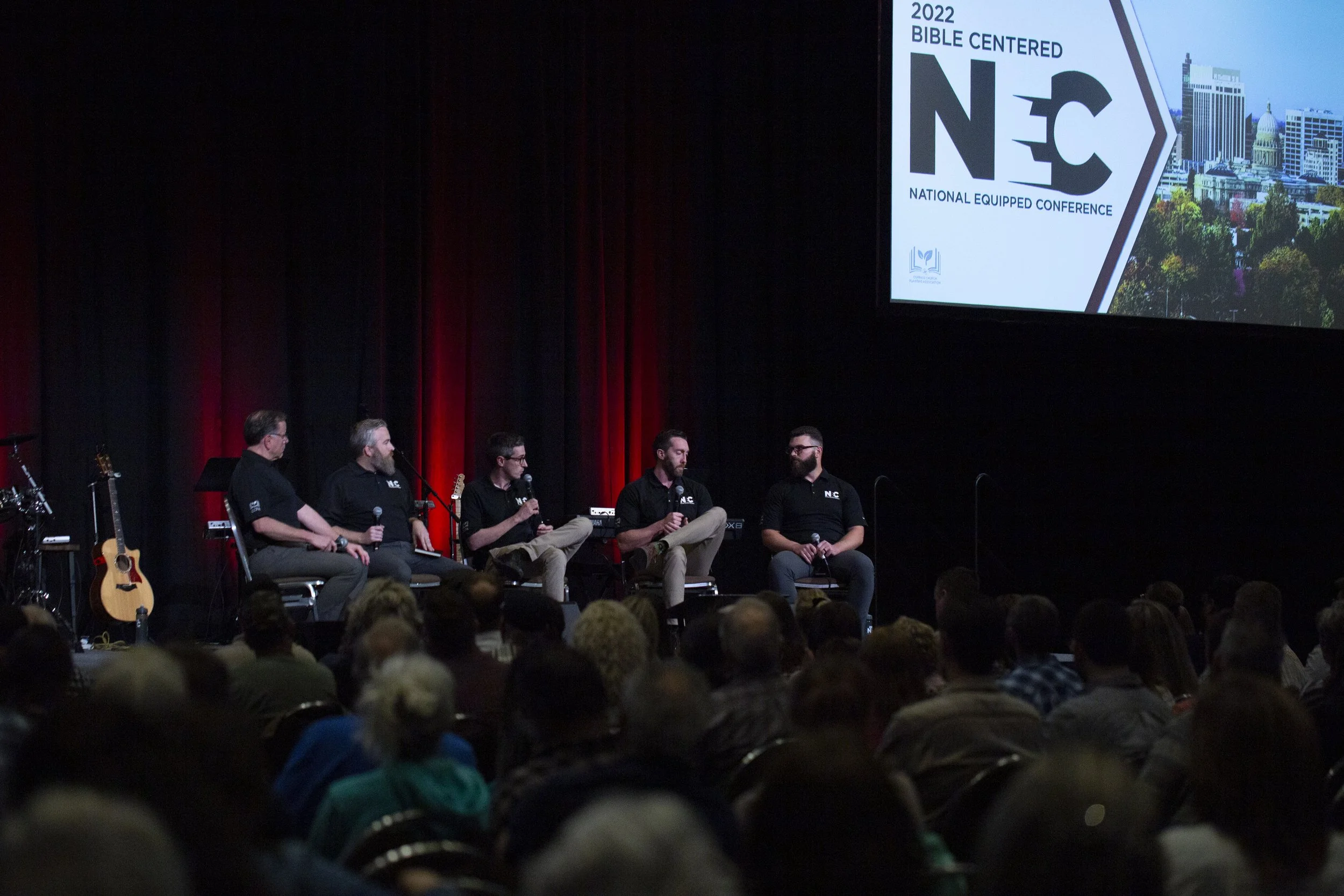 Five men are seated on stage at the 2022 Bible Centered NCC conference, speaking into microphones, with a large screen above them displaying the conference logo and a cityscape background, and a musical setup with guitars on the left.