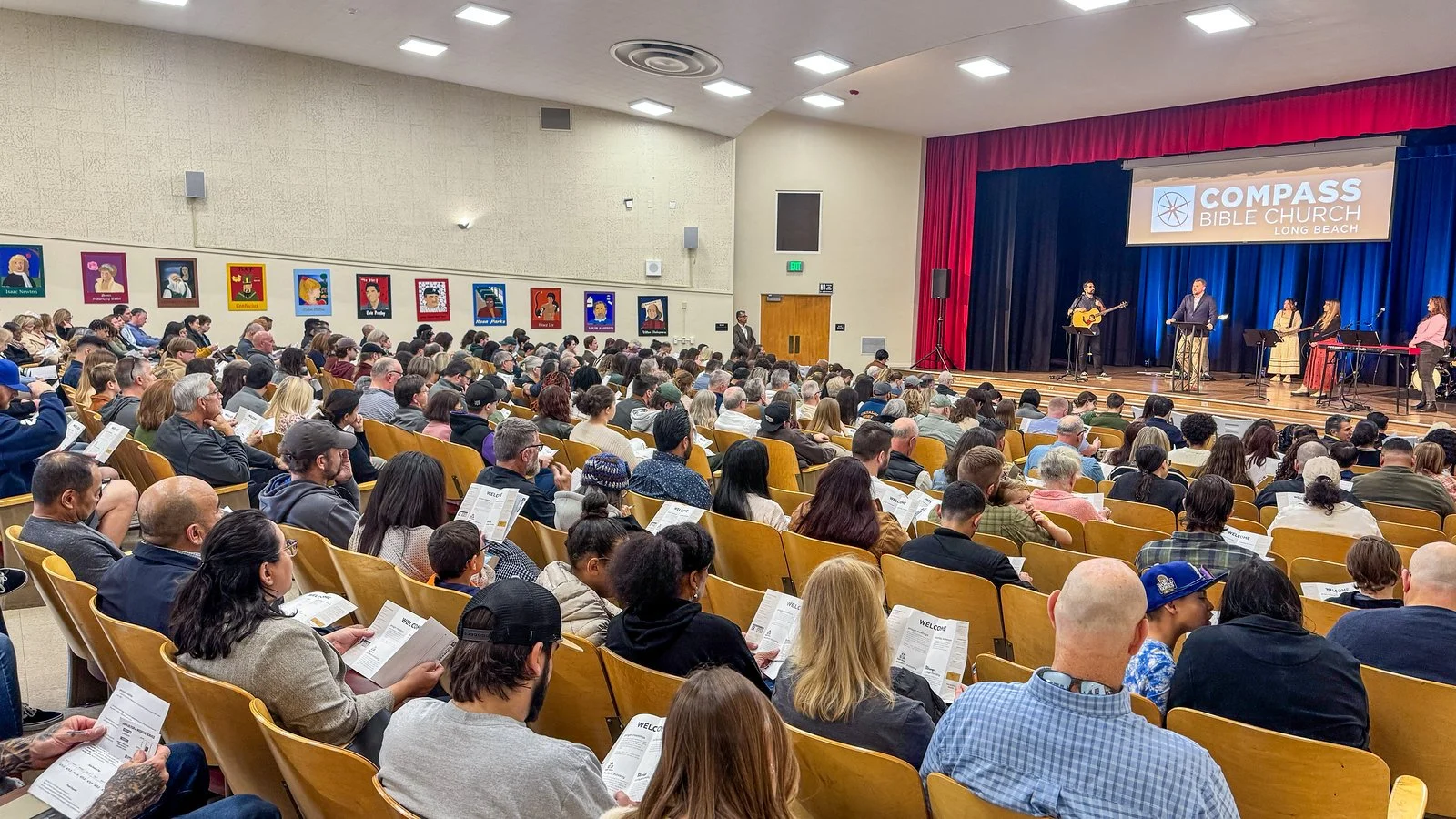 A large congregation seated in an auditorium with a stage at the front. The stage has musicians and a singer, with a large screen displaying the words 'COMPASS BIBLE CHURCH LONG BEACH.' The audience is engaged, some reading programs or pamphlets, and colorful artwork lines the wall on the left side.
