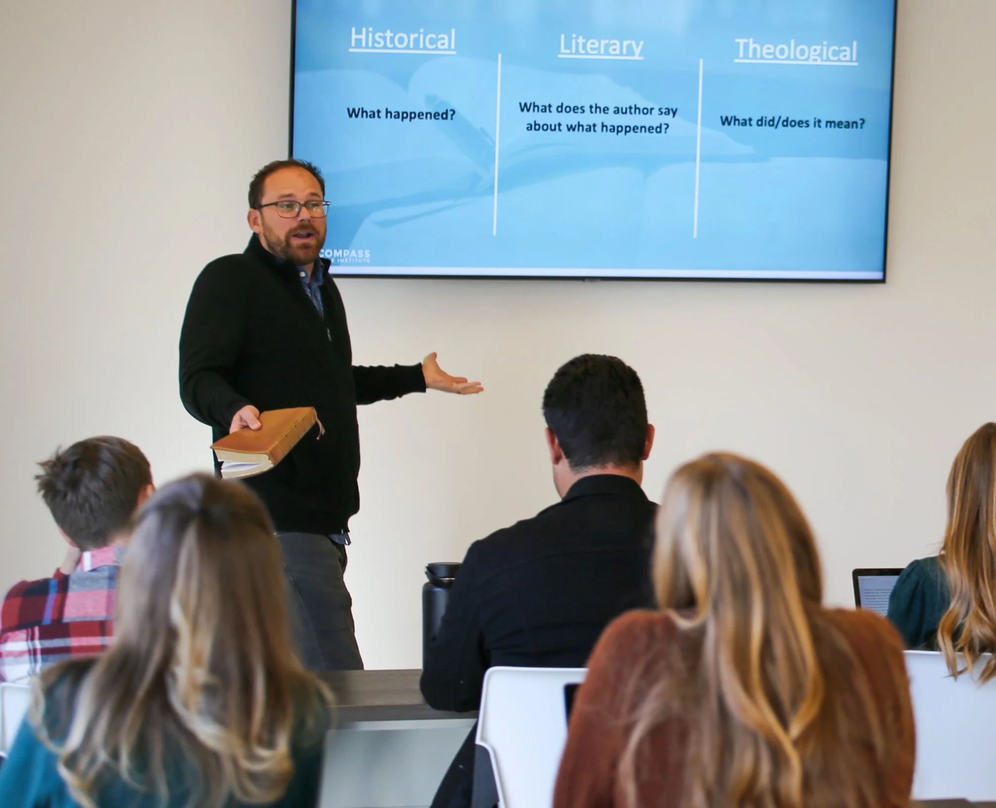 A man giving a lecture in front of a large screen displaying a chart with categories 'Historical,' 'Literary,' and 'Theological,' with descriptions under each. Several students are seated facing him, some with laptops.