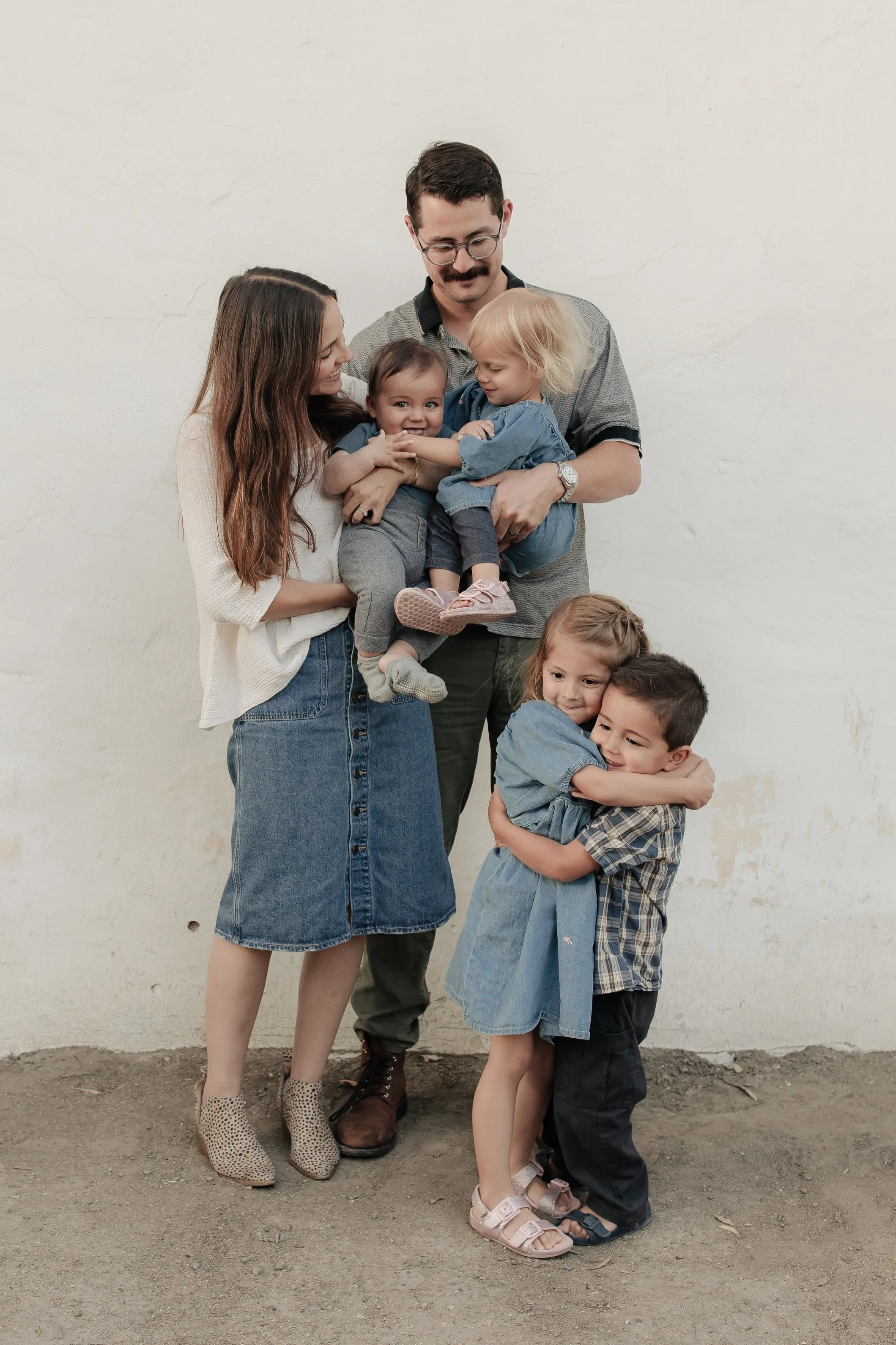 A family of five poses against a plain wall, with the parents holding their young children. The father, with glasses and a mustache, stands in the center, holding two children, one in each arm. The mother, with long brown hair, stands to his left, smiling at the children. The children are two girls and a boy, all dressed casually, with the oldest girl standing in front of the parents, while the youngest girl hugs the boy. All are smiling and appear joyful.