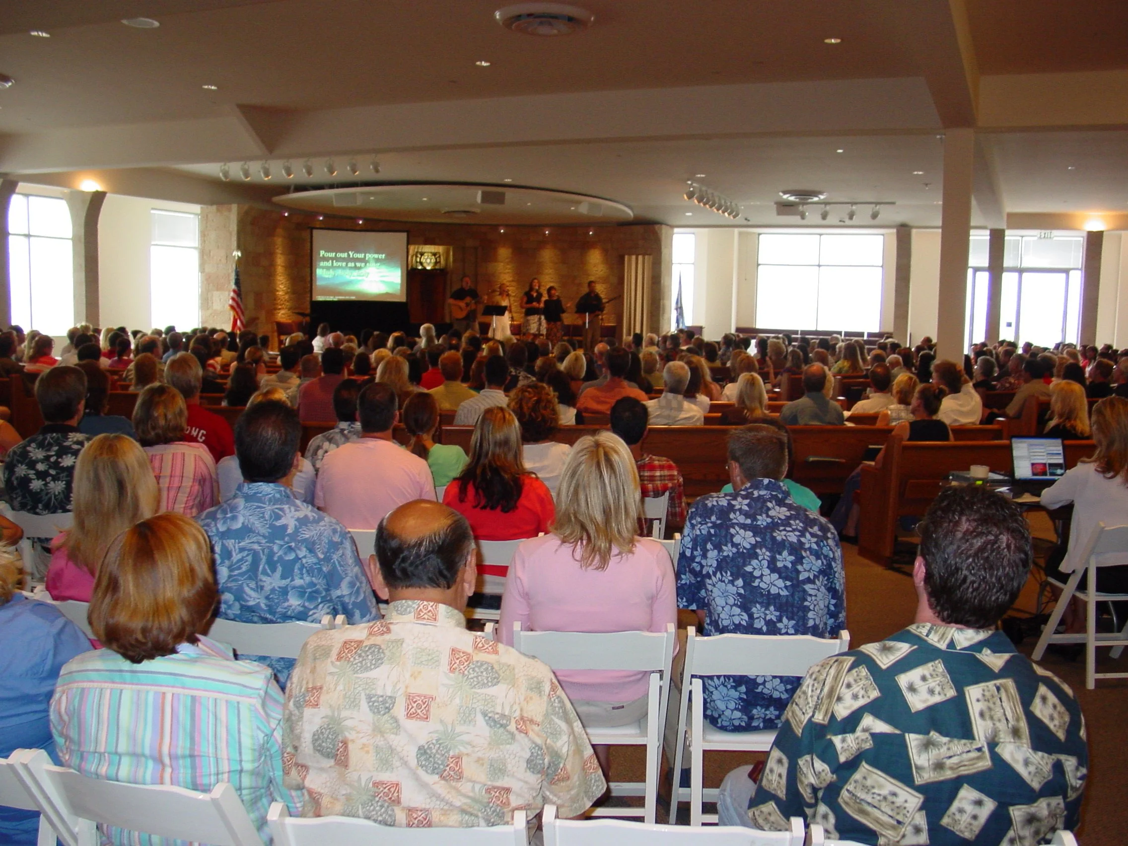 Large indoor church gathering with people seated in rows facing a stage, where a band is performing and lyrics are displayed on a screen.