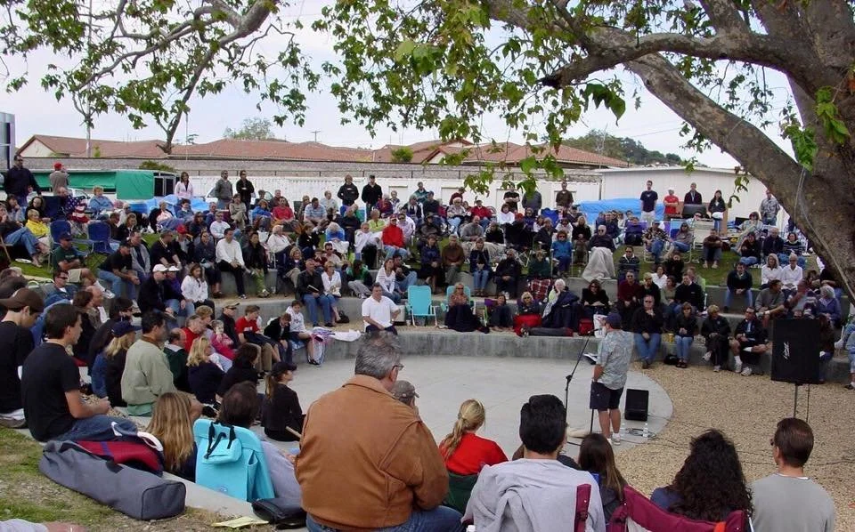 A large outdoor gathering with many people seated and standing, watching a speaker at a microphone in an amphitheater-like setting, with a tree overhead and buildings in the background.