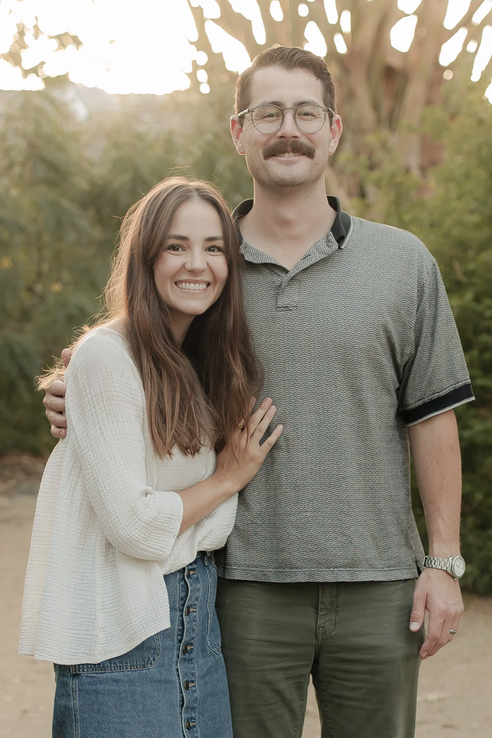 A smiling young woman with long brown hair and a man with glasses and a mustache pose together outdoors, with trees and sunlight in the background.