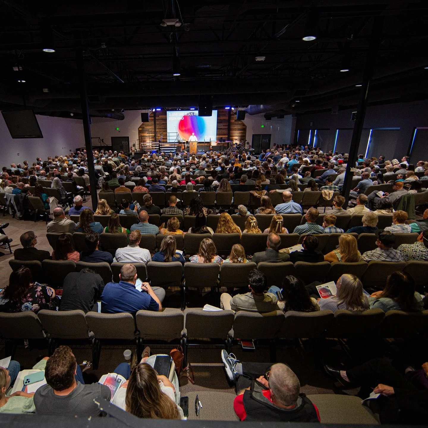 A large indoor gathering with numerous people seated in rows, watching a speaker on stage at a conference or church event.