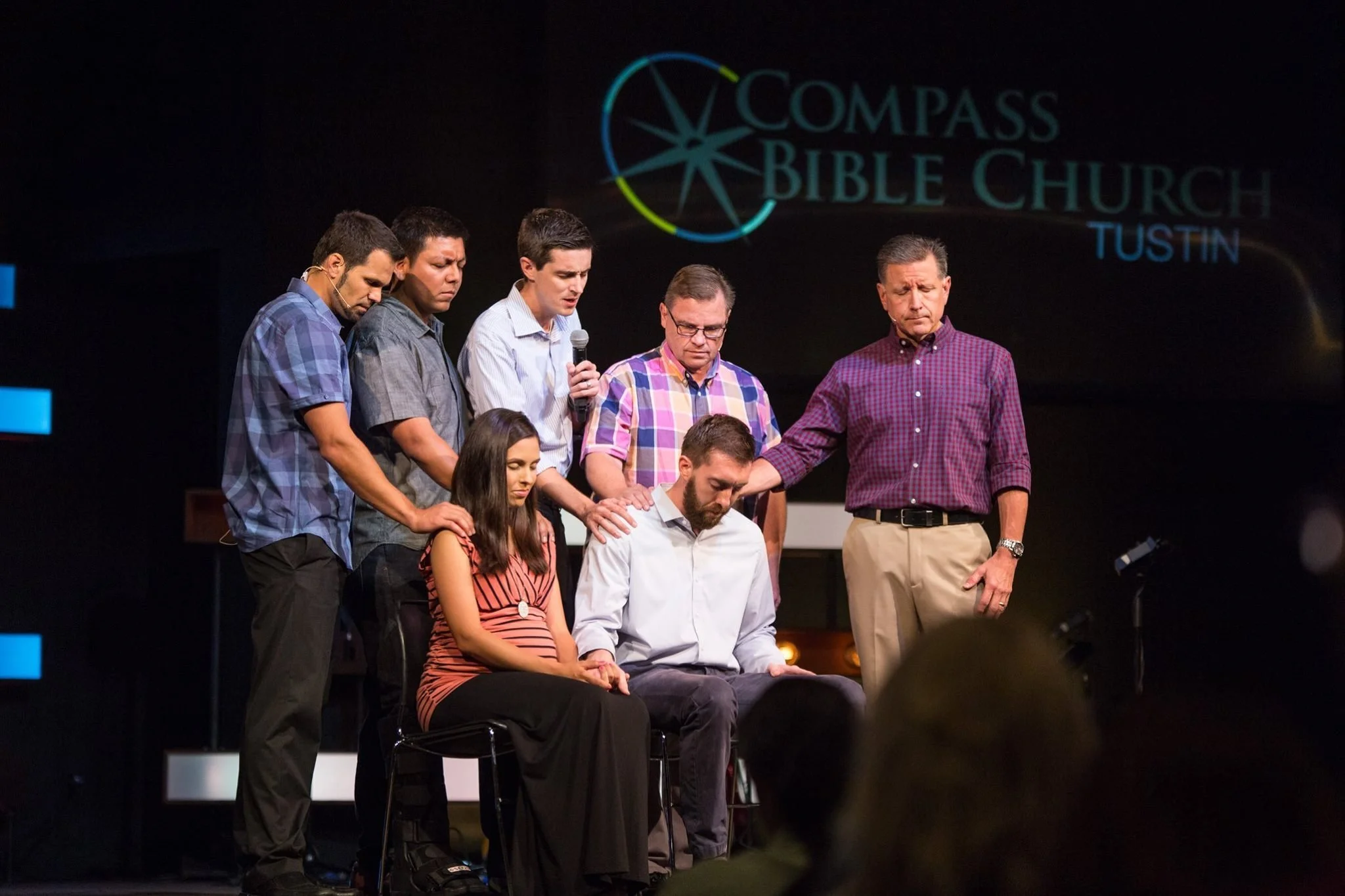 A group of people praying with their eyes closed and hands on a person sitting in front of them inside a church auditorium.
