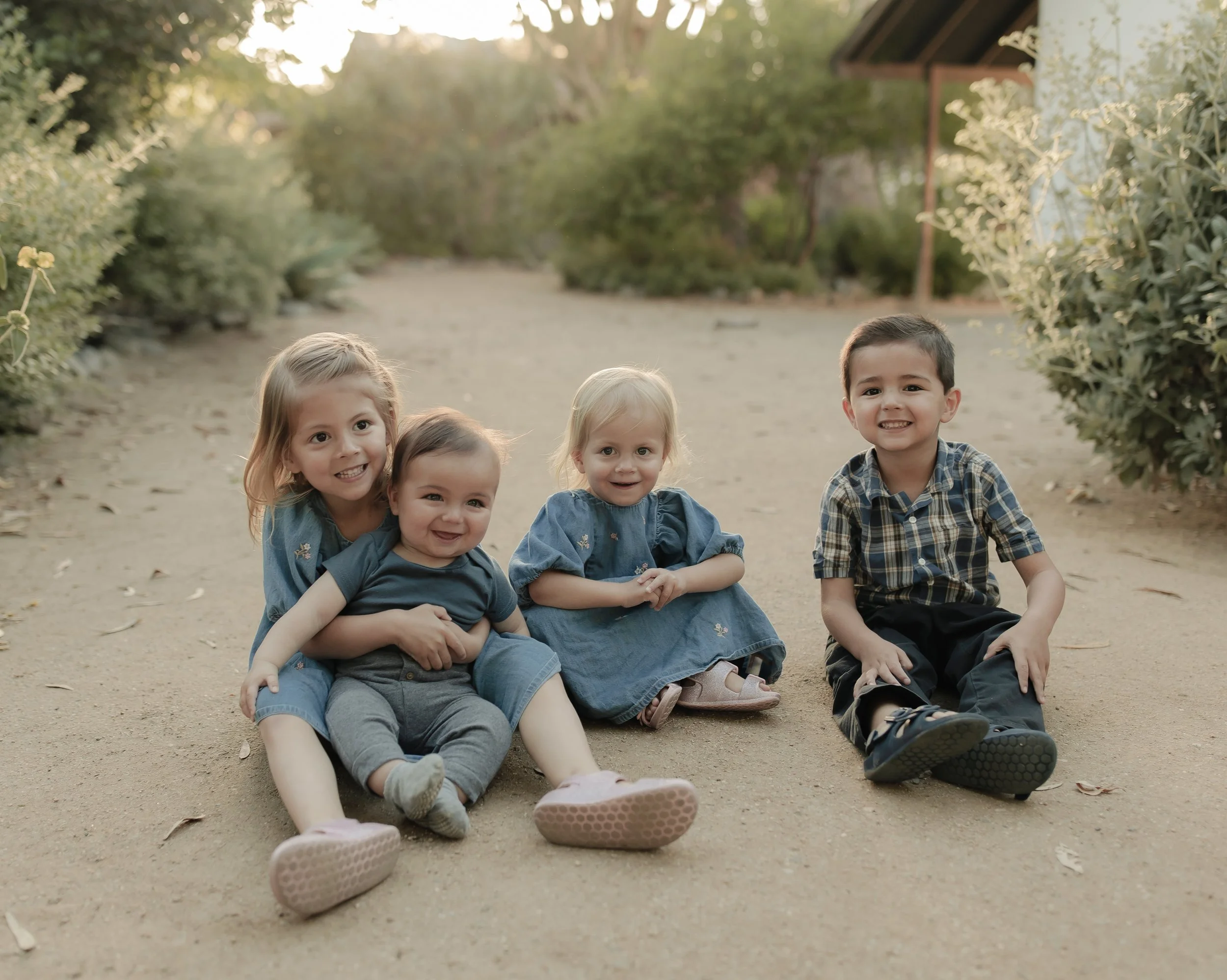 A group of five young children sitting together outdoors on a dirt path, smiling at the camera with greenery and trees in the background during late afternoon or early evening.
