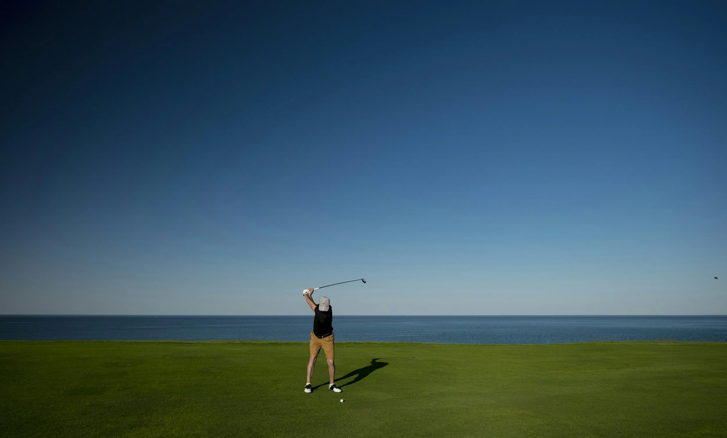 Un joueur de golf en position de swing se trouve sur un terrain de golf face à la mer, avec un ciel bleu dégagé et un oiseau volant au loin.