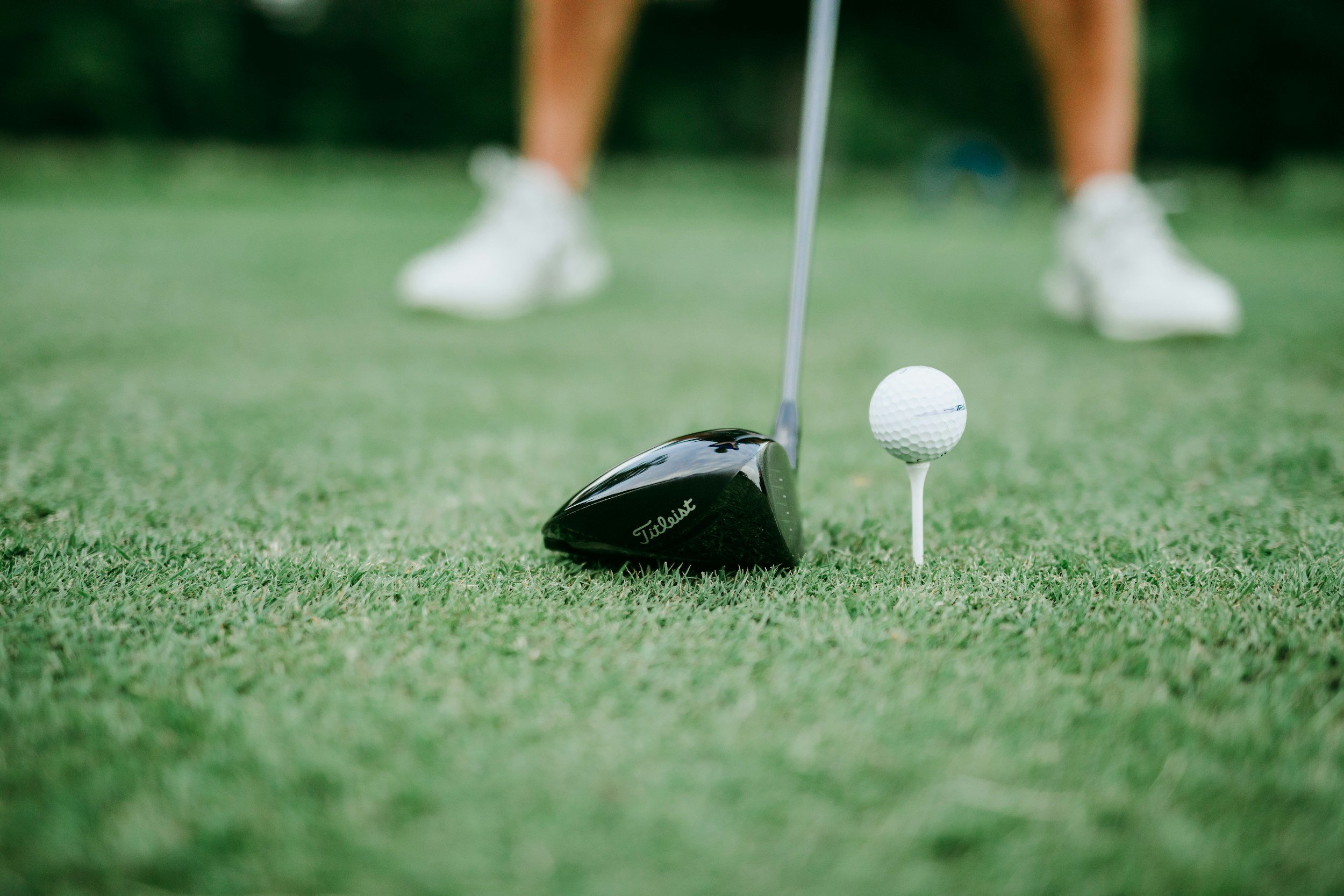 Une balle de golf blanche posée sur un tee blanc sur un terrain de golf vert, avec un club de golf noir appuyé sur le tee et deux personnes en arrière-plan en train de jouer au golf, portant des shorts et des chaussures de sport.